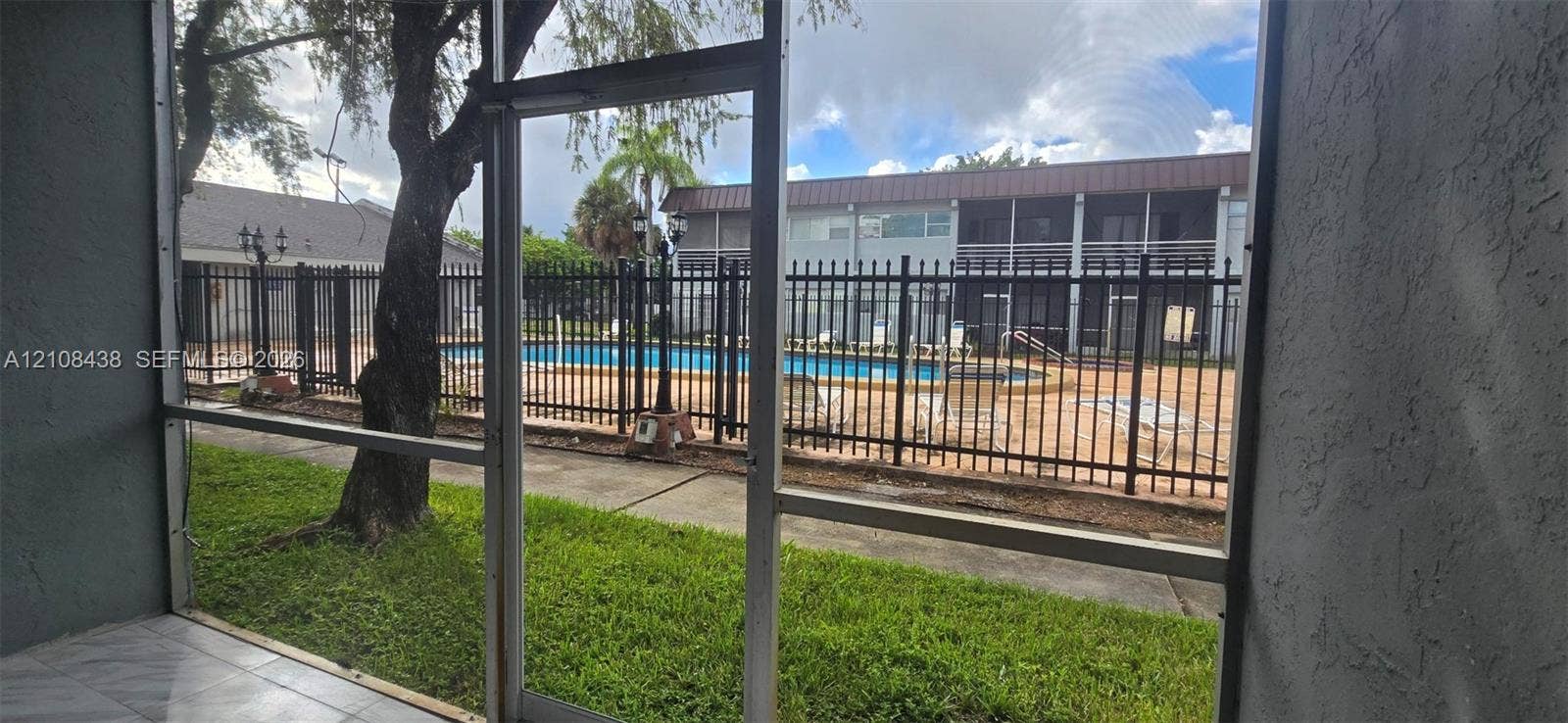 7320 NW 85th Court, #102, Tamarac, FL 33321 Screened-in patio with tile flooring, featuring proximity to a community swimming pool enclosed by black metal fencing and mature landscaping
