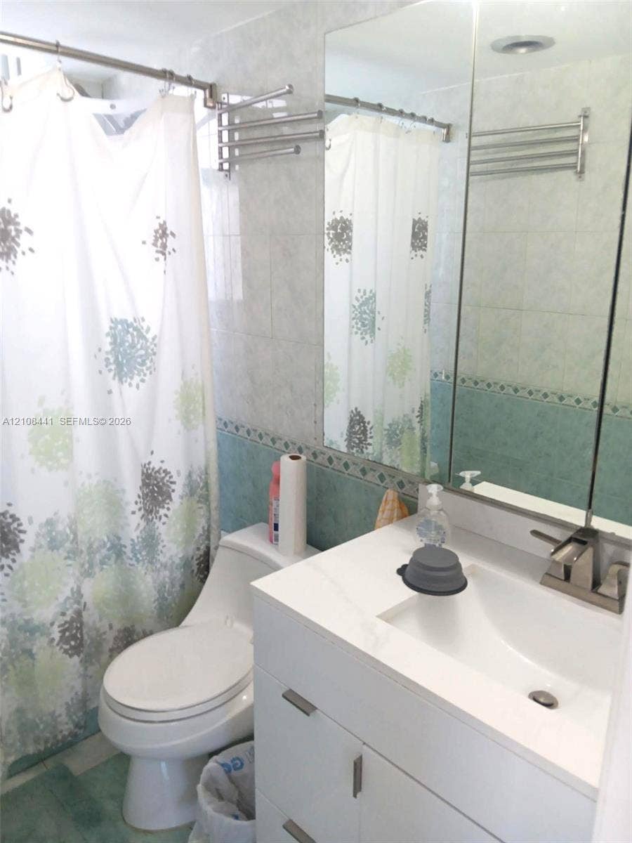 3703 NE 166th Street, #401, North Miami Beach, North Miami Beach, FL 33160 Bathroom featuring a white vanity cabinet with an integrated rectangular sink, a stone-finish countertop, a wall-mounted mirrored medicine cabinet, and tiled walls with decorative trim