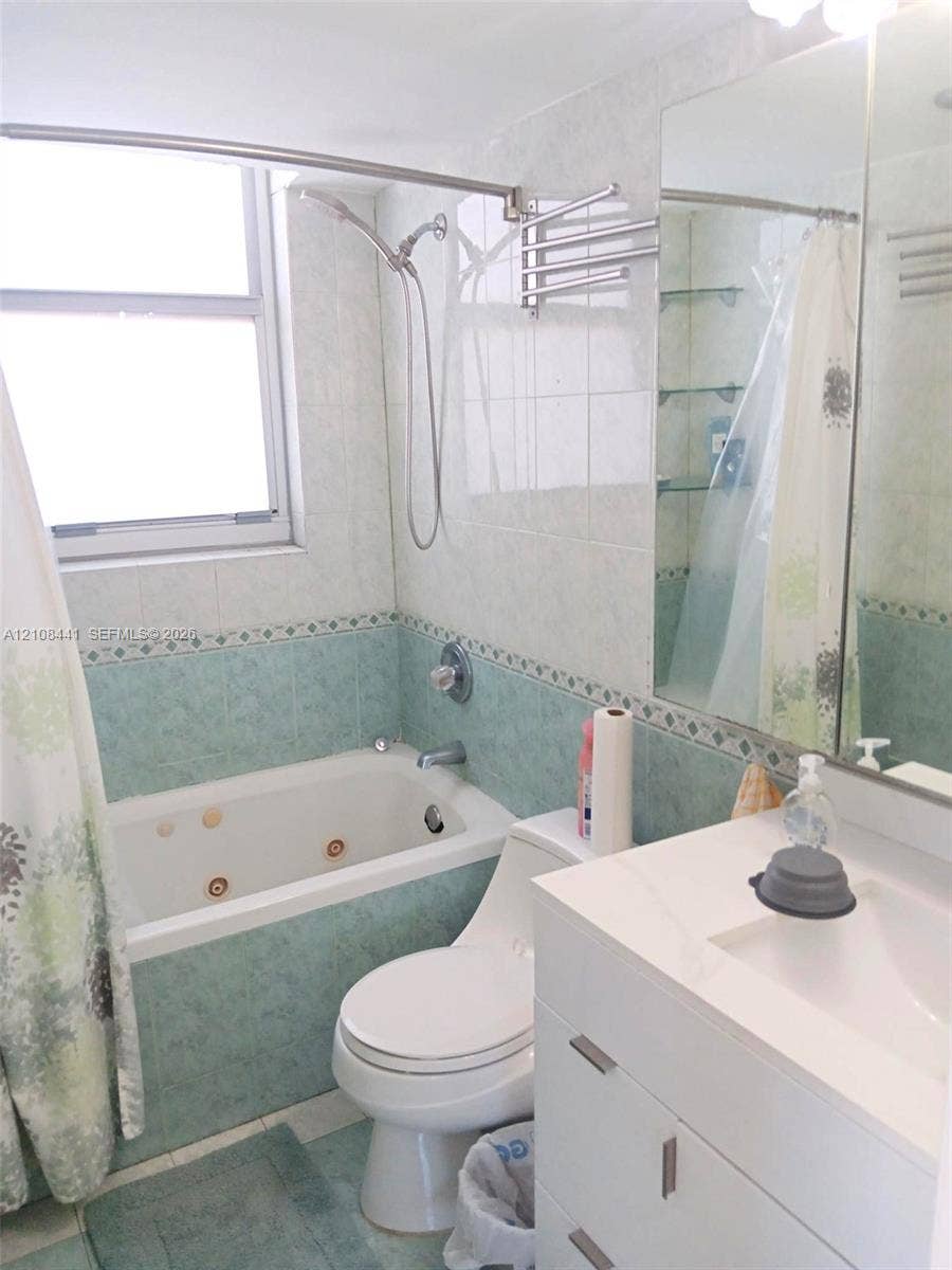 3703 NE 166th Street, #401, North Miami Beach, North Miami Beach, FL 33160 Bathroom featuring a jetted tub, wall-mounted handheld shower fixture, modern white vanity with a rectangular integrated sink, large vanity mirror, and glass wall shelving