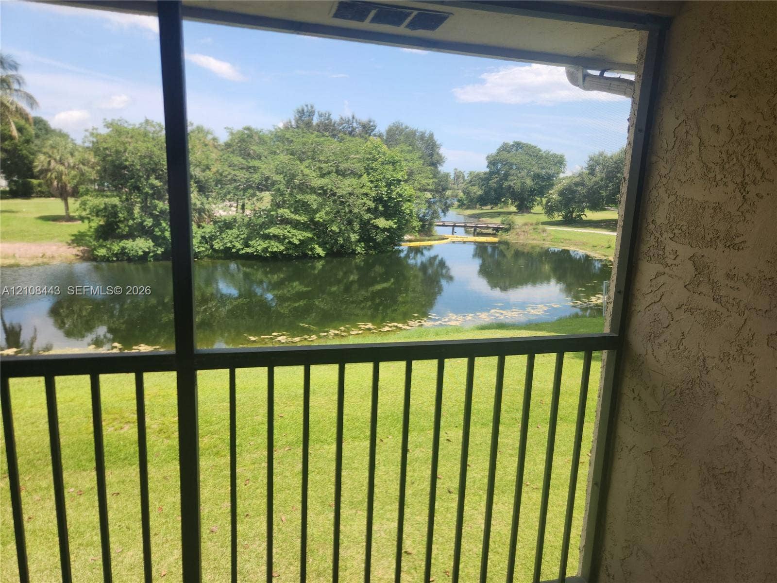 4018 NW 87th Avenue, #4018, Sunrise, Sunrise, FL 33351 Screened-in balcony with metal railing, overlooking a pond, mature trees, and pedestrian bridge