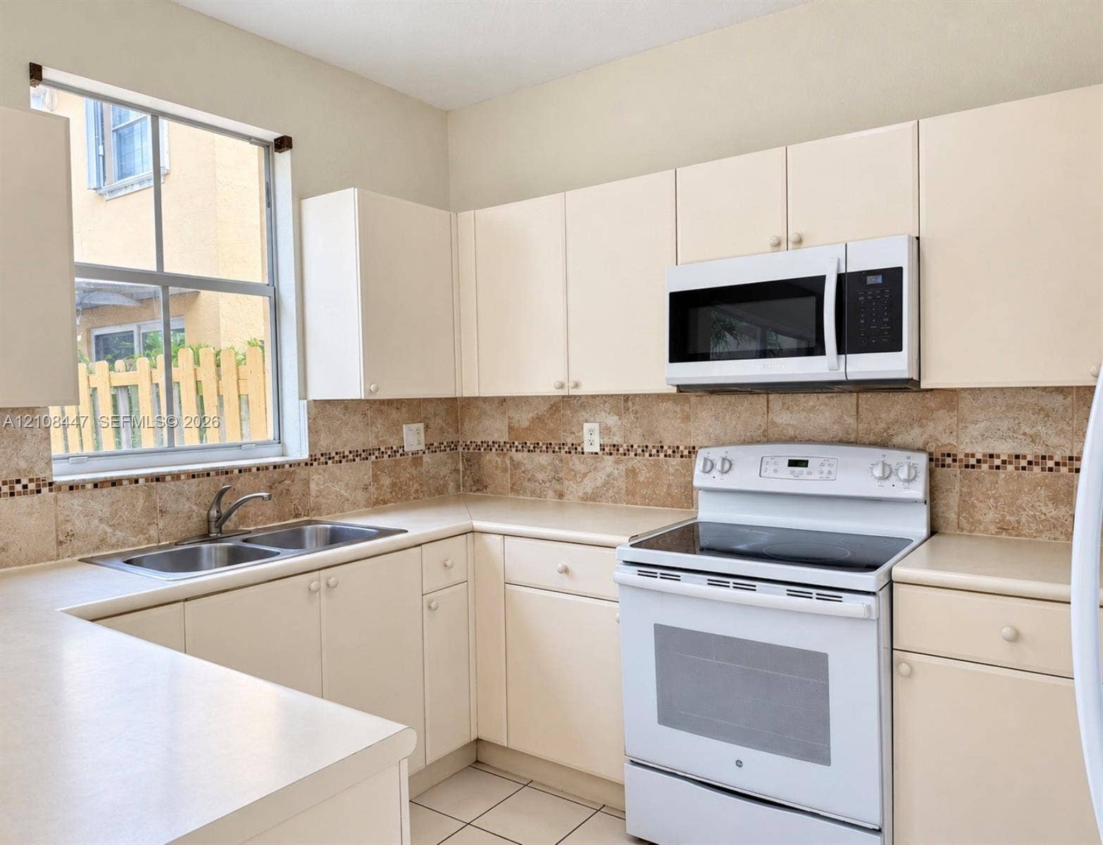 Kitchen featuring light-finish cabinetry, stone-texture tile backsplash with decorative inlay, solid-surface countertops, double-basin sink, and built-in microwave