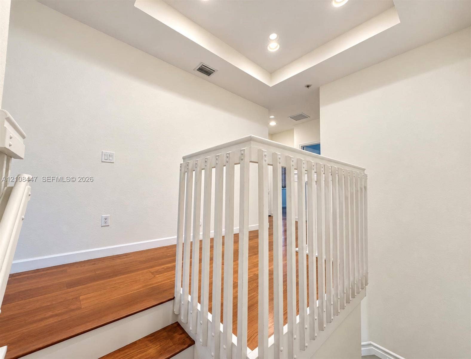 Recessed lighting, wood-finish flooring, and white vertical baluster railing surrounding the stairwell with a tray ceiling detail