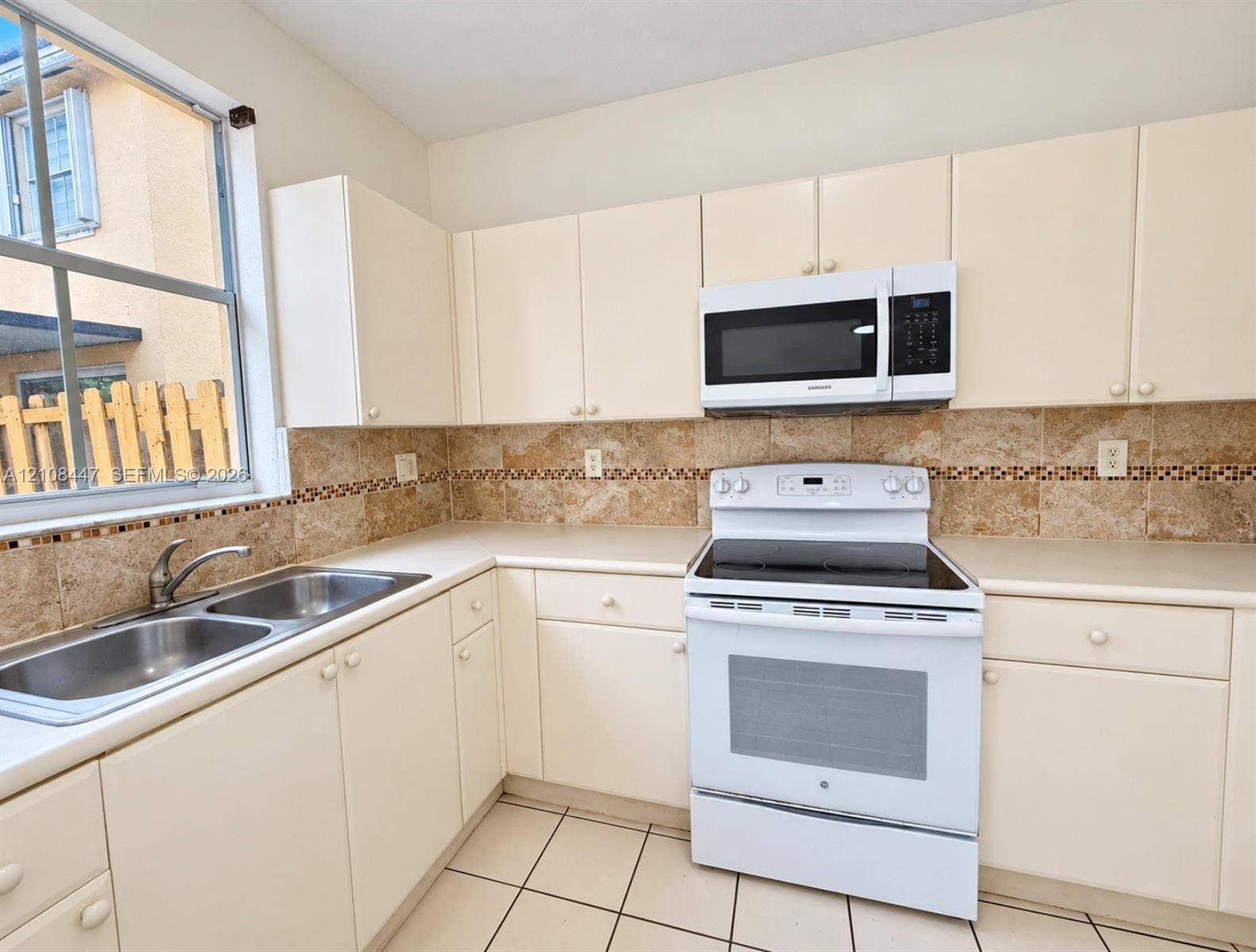 Kitchen featuring off-white cabinetry, stone-tiled backsplash with decorative inlay, solid surface countertops, dual-basin stainless steel sink with pull-out faucet, and white appliances including an electric range and built-in microwave