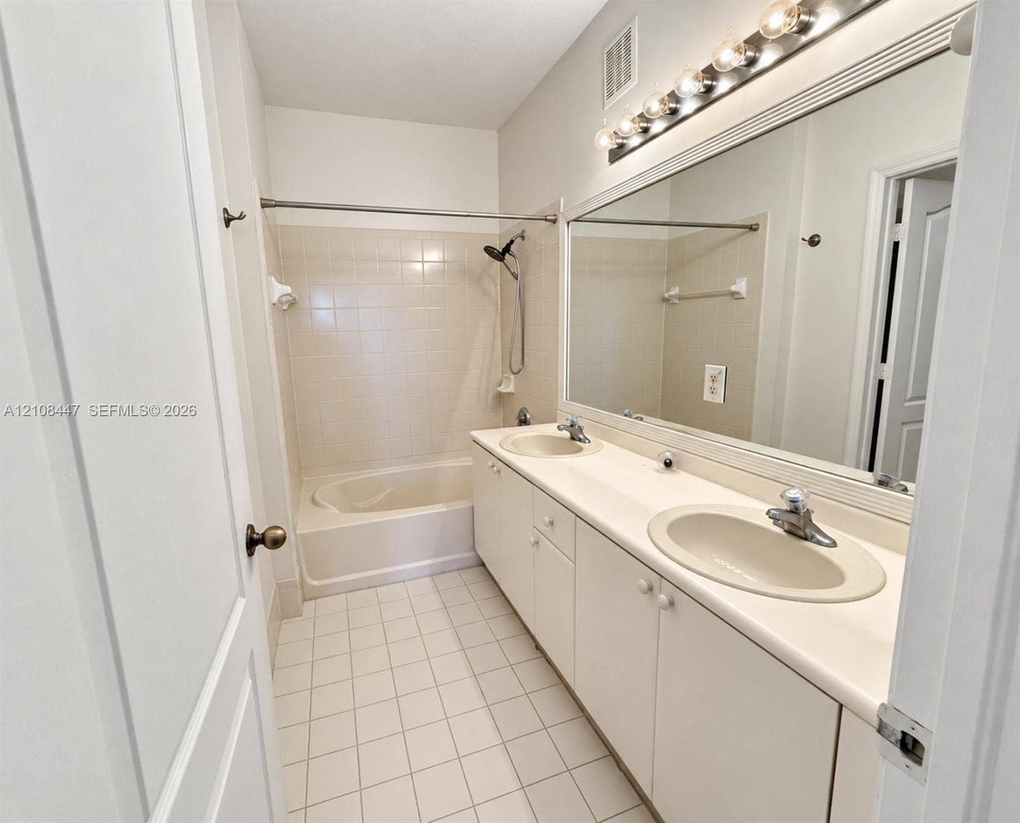 Bathroom featuring a dual-sink vanity with a solid-surface countertop, full-width vanity mirror, tile flooring, and a combined tub and shower enclosure with tile surround
