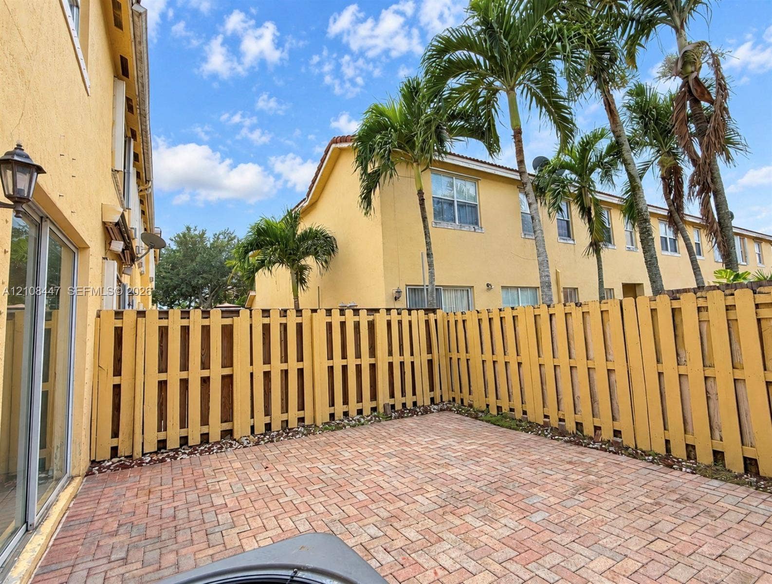Private patio featuring herringbone-patterned brick pavers and a wooden privacy fence, complemented by mature palm trees