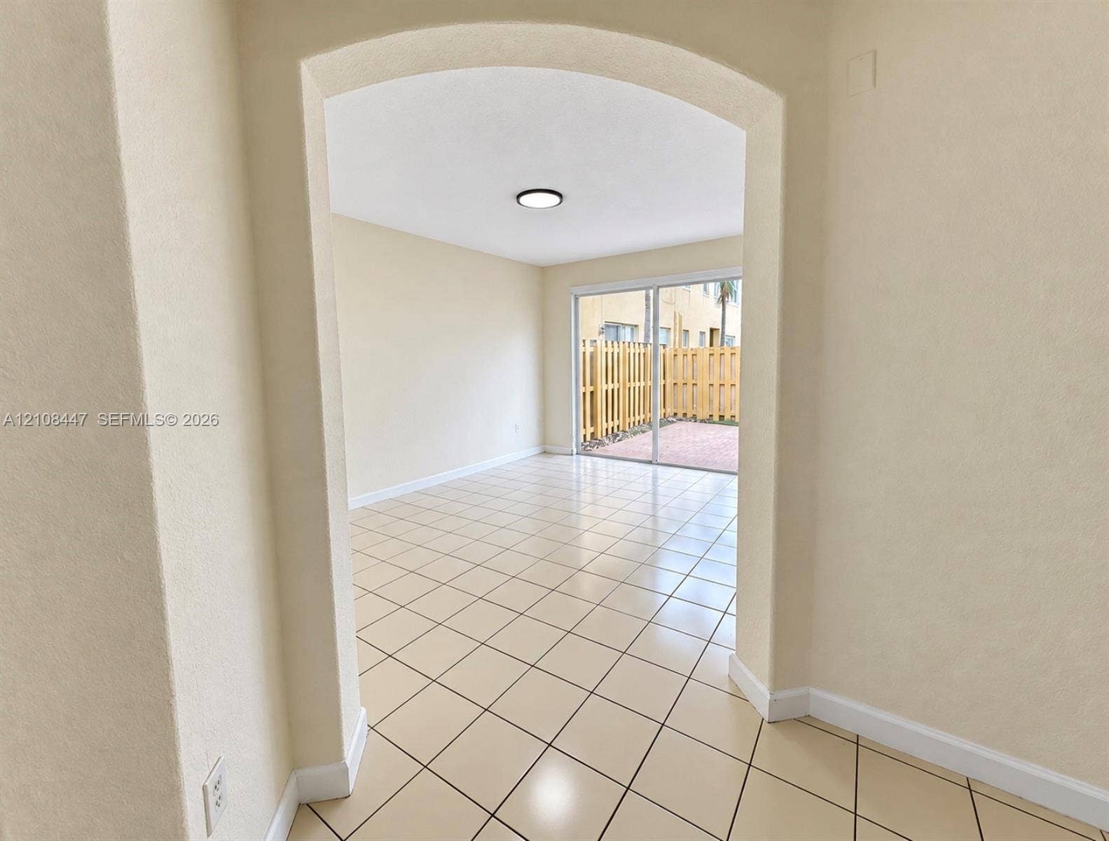 Arched entryway leading to a room with neutral ceramic tile flooring, flush-mount lighting, and glass sliding doors opening to a paved outdoor area with wood-style fencing