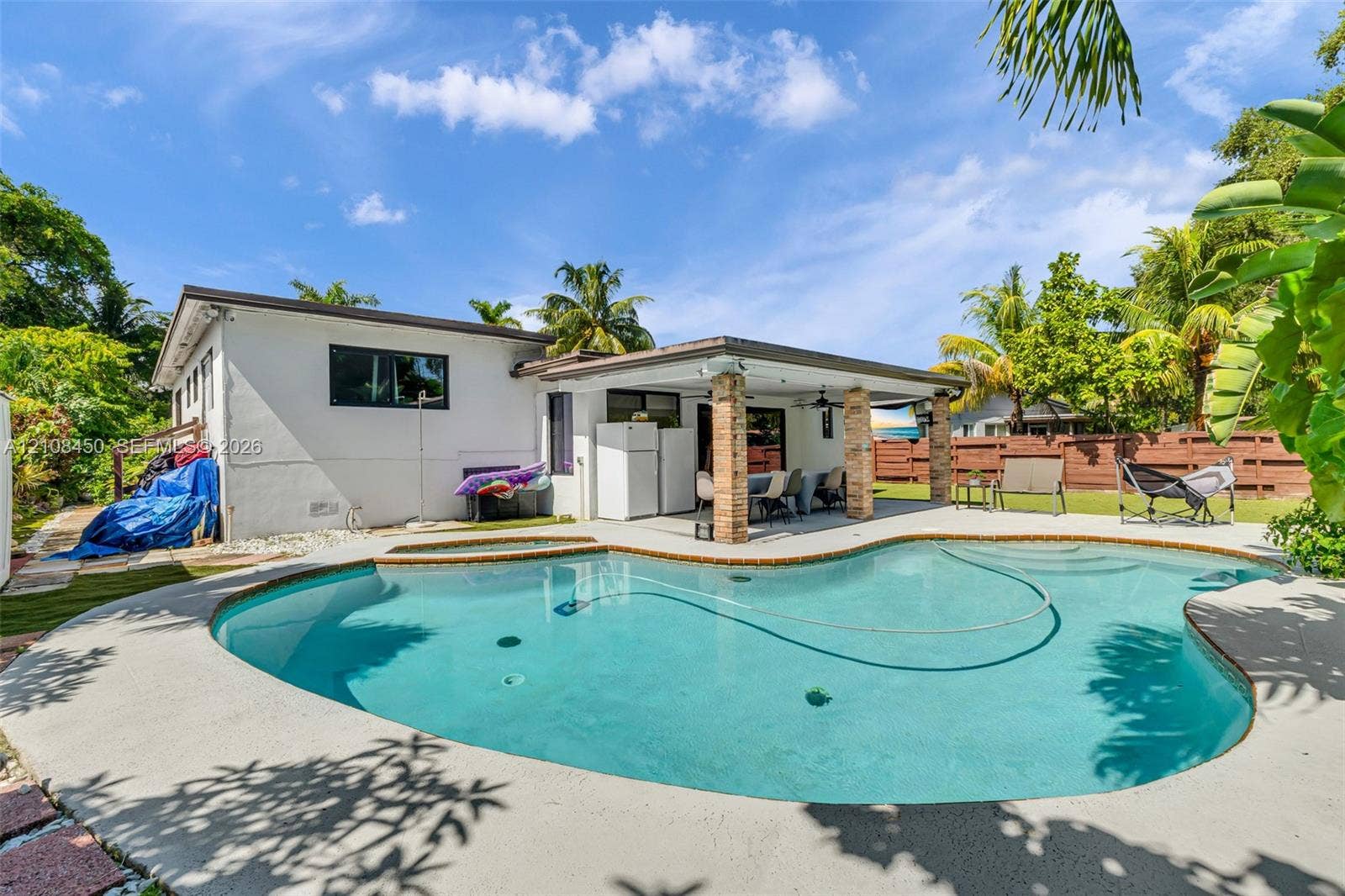 In-ground swimming pool with stone-tiled spa, concrete pool deck, covered patio with stone-finished support columns, and perimeter wood fencing