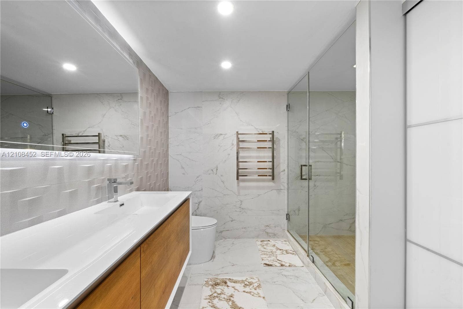 1000 Parkview Drive, #809, Hallandale Beach, FL 33009 Floating wood-finish vanity with an integrated rectangular sink, wall-mounted metallic faucet, large frameless vanity mirror, textured three-dimensional wall tile, and glass-enclosed shower with floor-to-ceiling stone-look tile