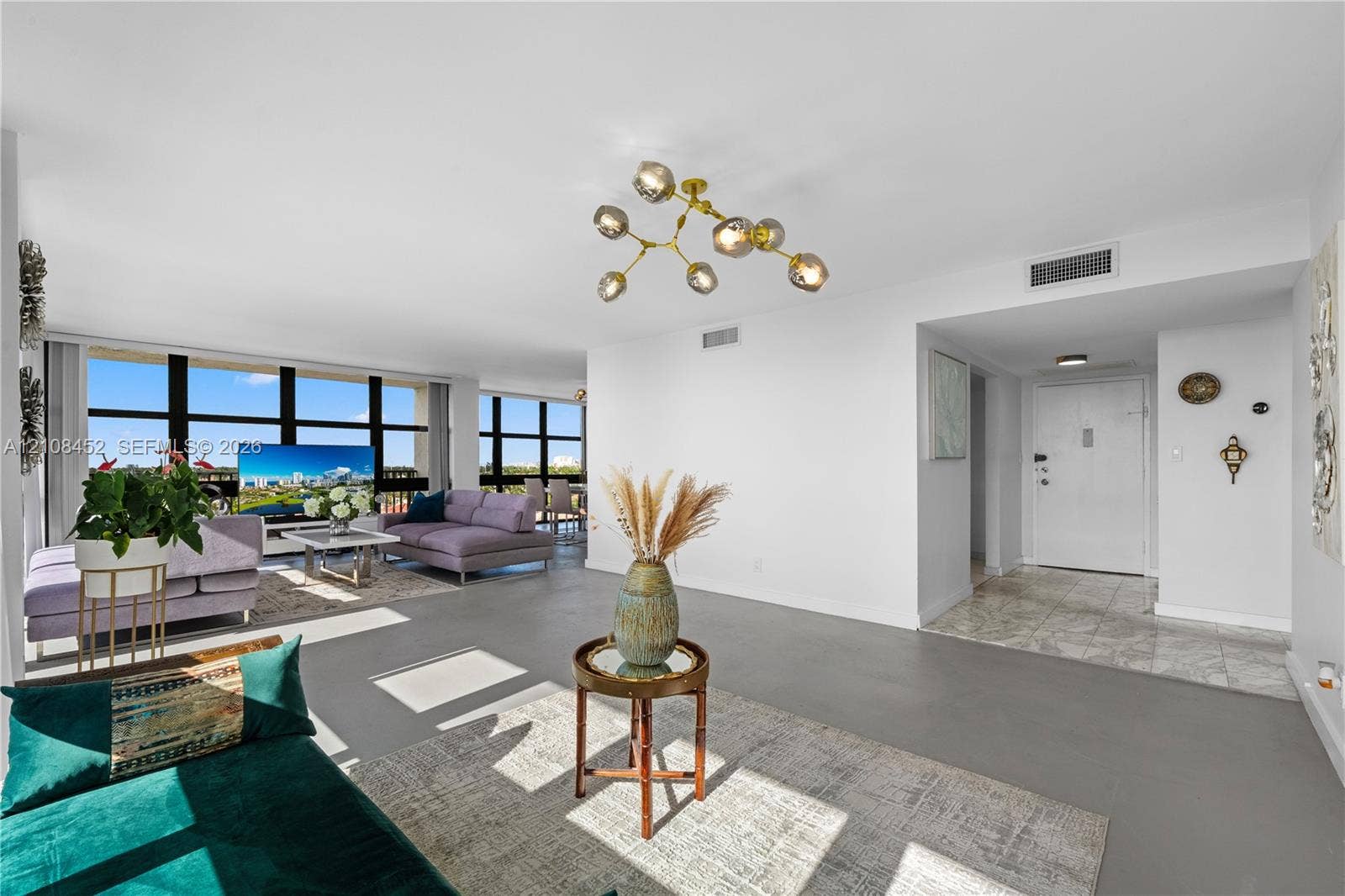 1000 Parkview Drive, #809, Hallandale Beach, FL 33009 Open-concept living space featuring floor-to-ceiling black-frame windows, neutral tone flooring, and a multi-light brass-finish chandelier