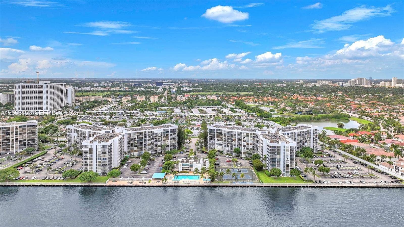 1000 Parkview Drive, #809, Hallandale Beach, FL 33009 Aerial perspective of waterfront residential complex featuring expansive surface parking, on-site swimming pool, hard-surfaced tennis courts, and mature landscaping