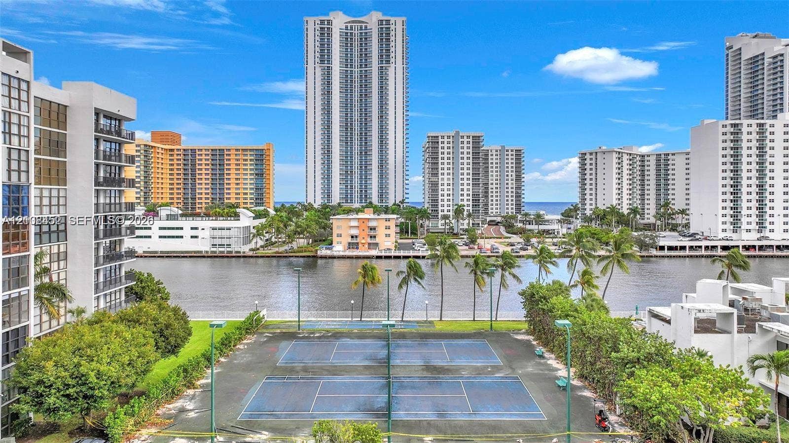 1000 Parkview Drive, #809, Hallandale Beach, FL 33009 Outdoor tennis court, waterfront setting, paved perimeter walking path, manicured lawn areas, and mature palms