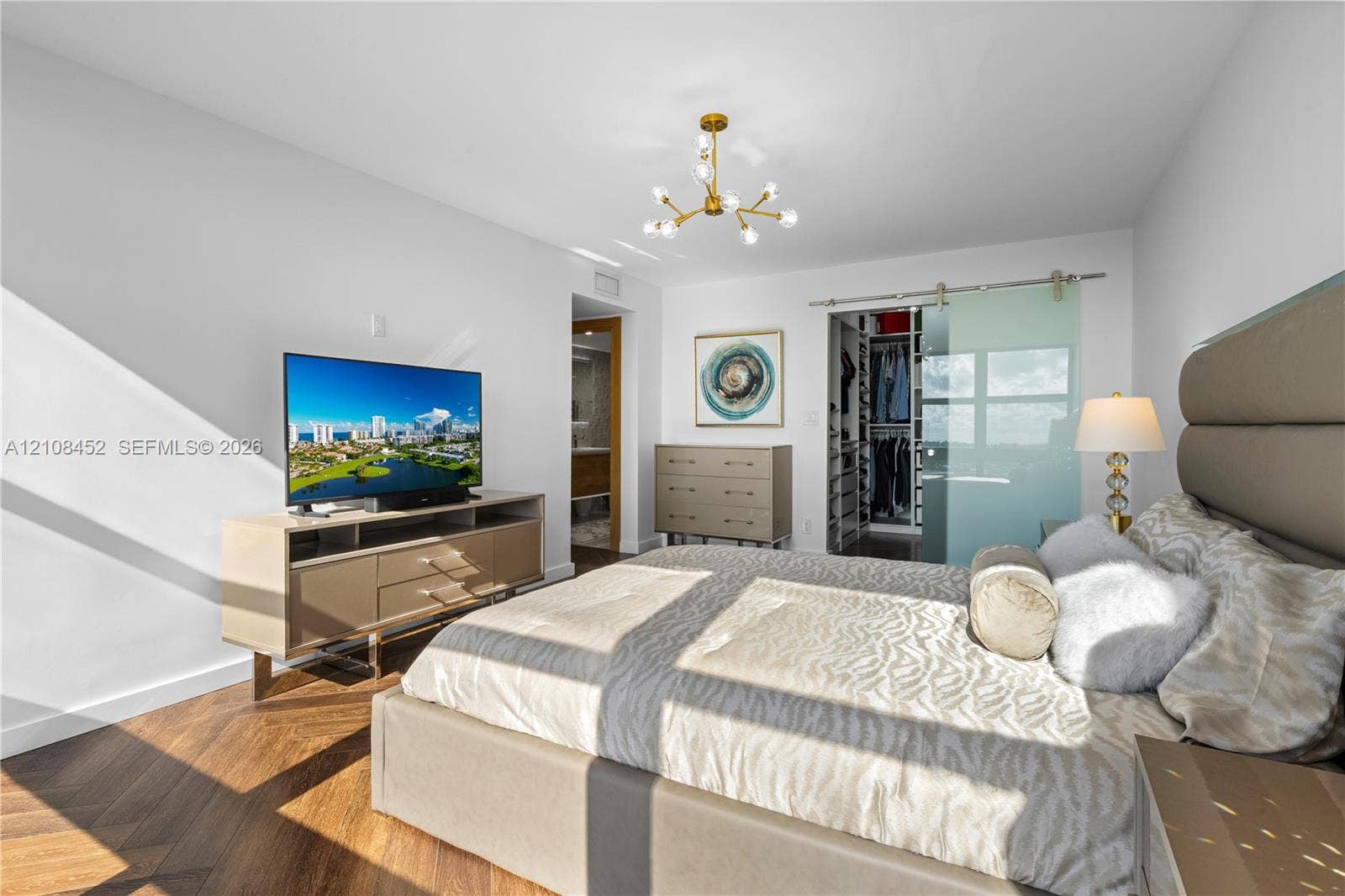 1000 Parkview Drive, #809, Hallandale Beach, FL 33009 Herringbone-pattern wood-finish flooring, gold-tone modern multi-arm chandelier, and frosted glass sliding barn-style door opening to a walk-in closet
