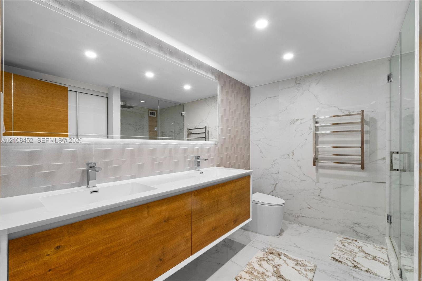 1000 Parkview Drive, #809, Hallandale Beach, FL 33009 Floating wood-finish double vanity with rectangular integrated sinks, full-width mirror, recessed ceiling lighting, glass-enclosed shower, and heated towel rack