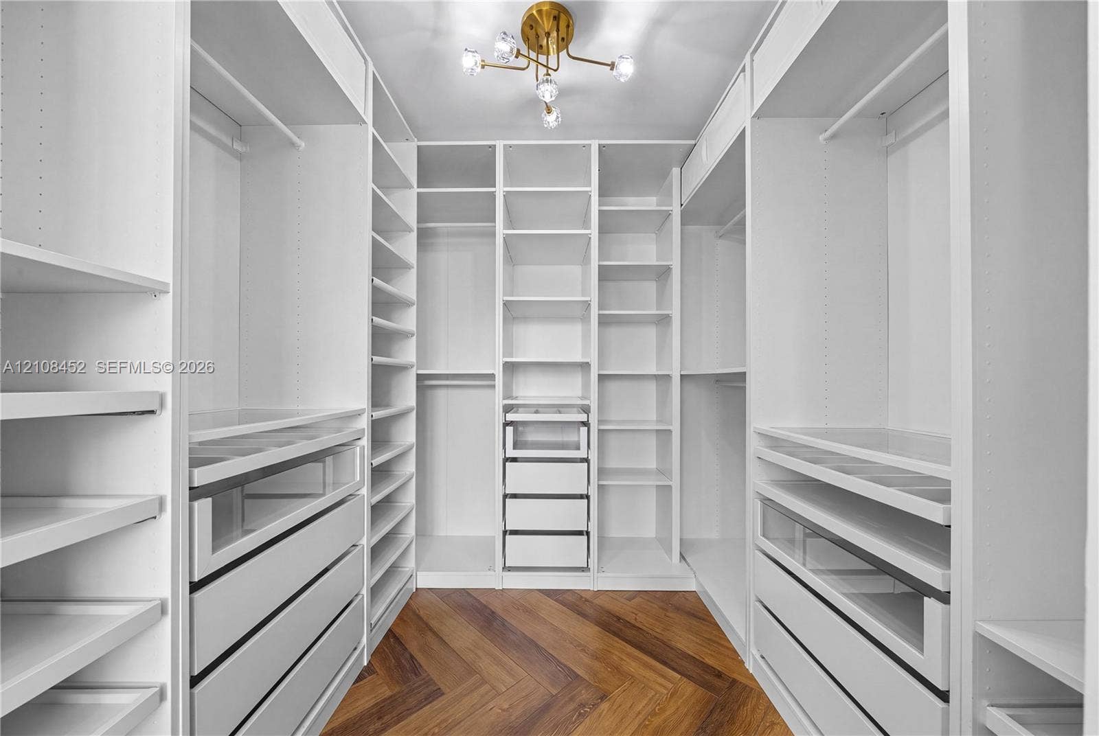 1000 Parkview Drive, #809, Hallandale Beach, FL 33009 Walk-in closet featuring extensive built-in white cabinetry, multiple shelving tiers, integrated drawer units with glass-front detailing, chevron-patterned wood-finish flooring, and a multi-arm metallic light fixture