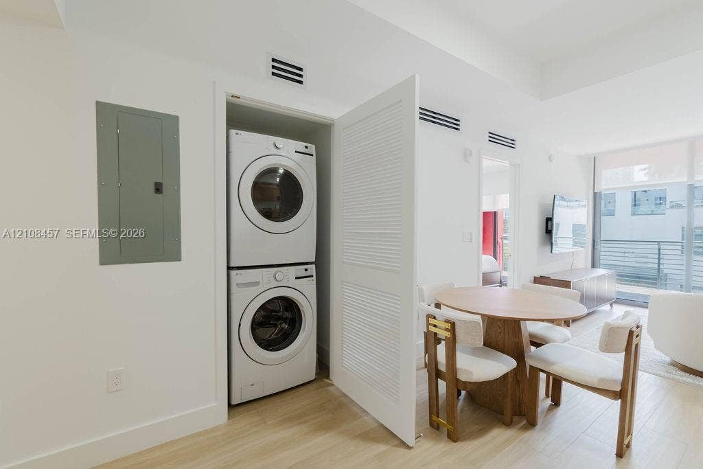 90 NE 32nd Street, #511, Miami, FL 33127 Stacked laundry center featuring a high-efficiency front-load washer and dryer enclosed by a louvered door, wood-finish flooring, and minimalist white interior walls