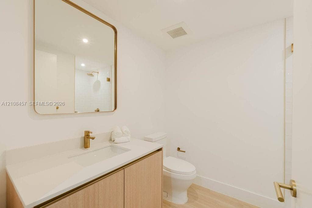 90 NE 32nd Street, #511, Miami, FL 33127 Modern vanity with wood-finish cabinetry, white countertop, integrated rectangular sink, brass-toned hardware, and a matching rounded-edge wall mirror