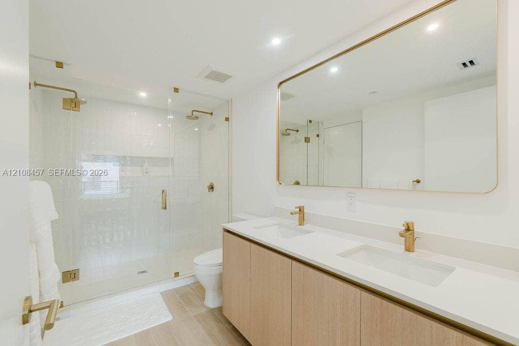 90 NE 32nd Street, #511, Miami, FL 33127 Double vanity with light wood-finish cabinetry, quartz countertop, dual undermount sinks, brass fixtures, and a glass-enclosed shower