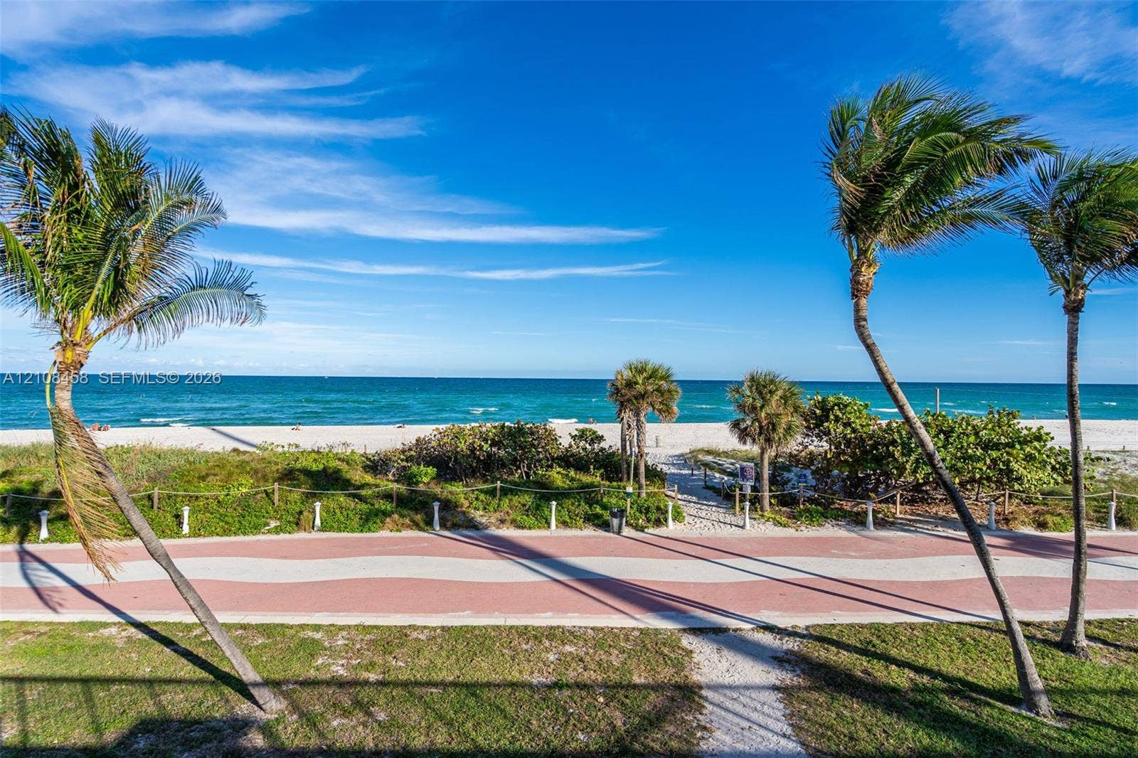 Coastal frontage featuring a paved pedestrian pathway, manicured landscaping, mature palm trees, and direct access to a sandy beach