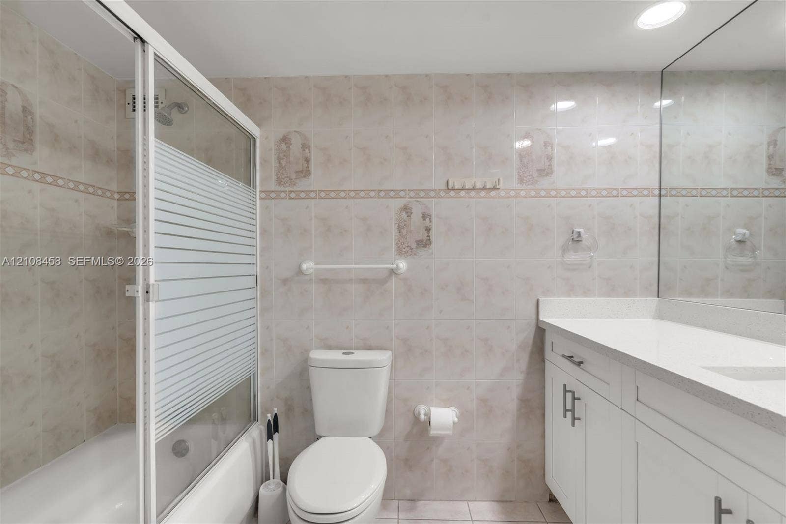Bathroom featuring white shaker-style cabinetry, quartz countertop with integrated basin, floor-to-ceiling tile walls with decorative accents, and a tub-shower combination with a sliding glass door