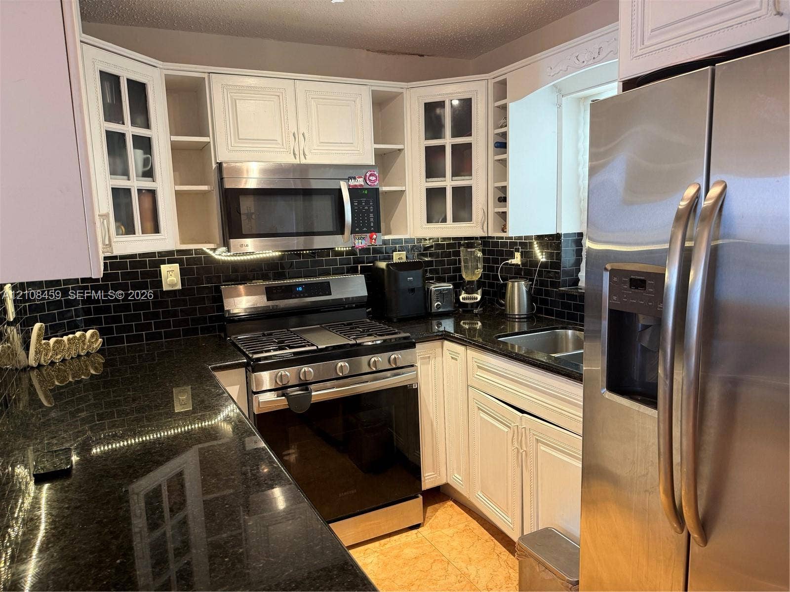 720 Collins Avenue, #206, Miami Beach, Miami Beach, FL 33139 Kitchen featuring white cabinetry with glass-panel display doors, stone countertops, black subway tile backsplash, and stainless steel appliances including a gas range and built-in microwave