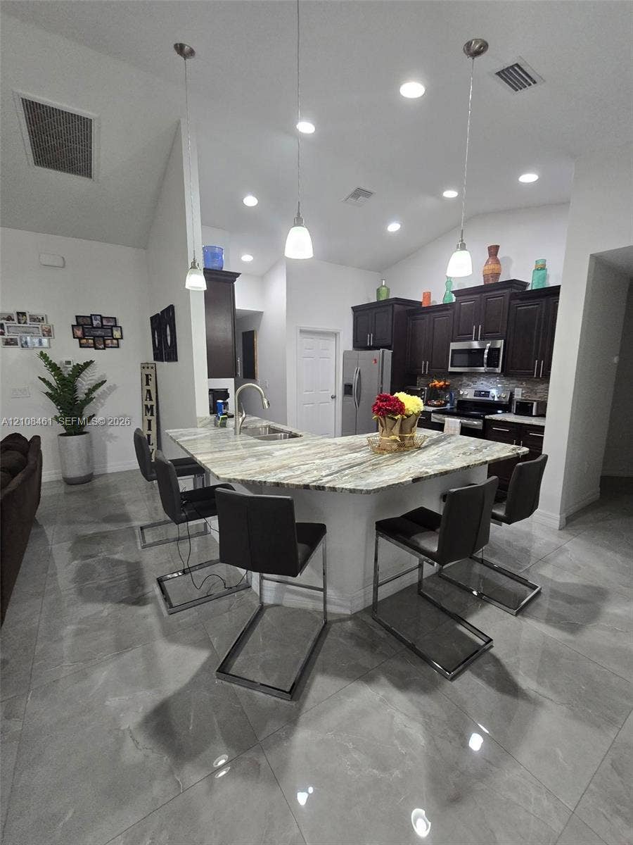 Kitchen featuring dark-finish cabinetry, stone countertops, a multi-seat island with an integrated stainless steel sink, tile flooring, and recessed ceiling lighting