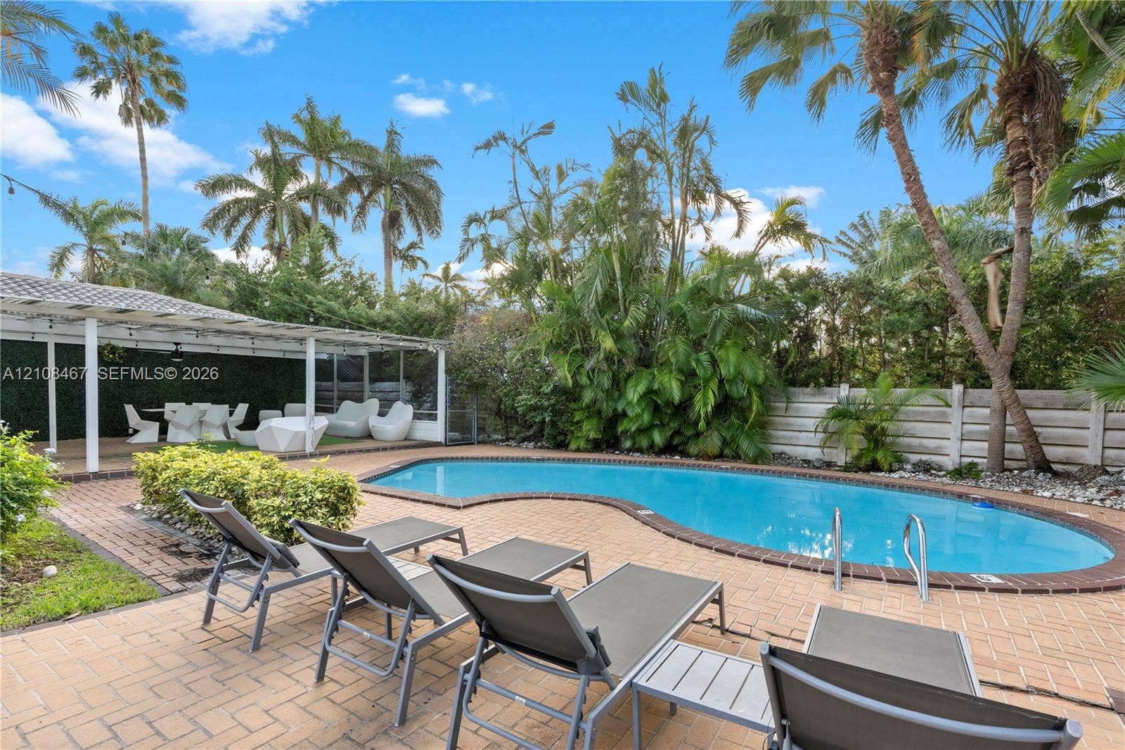 Freeform swimming pool featuring brick coping and a paved patio, complemented by a covered outdoor structure and mature tropical landscaping