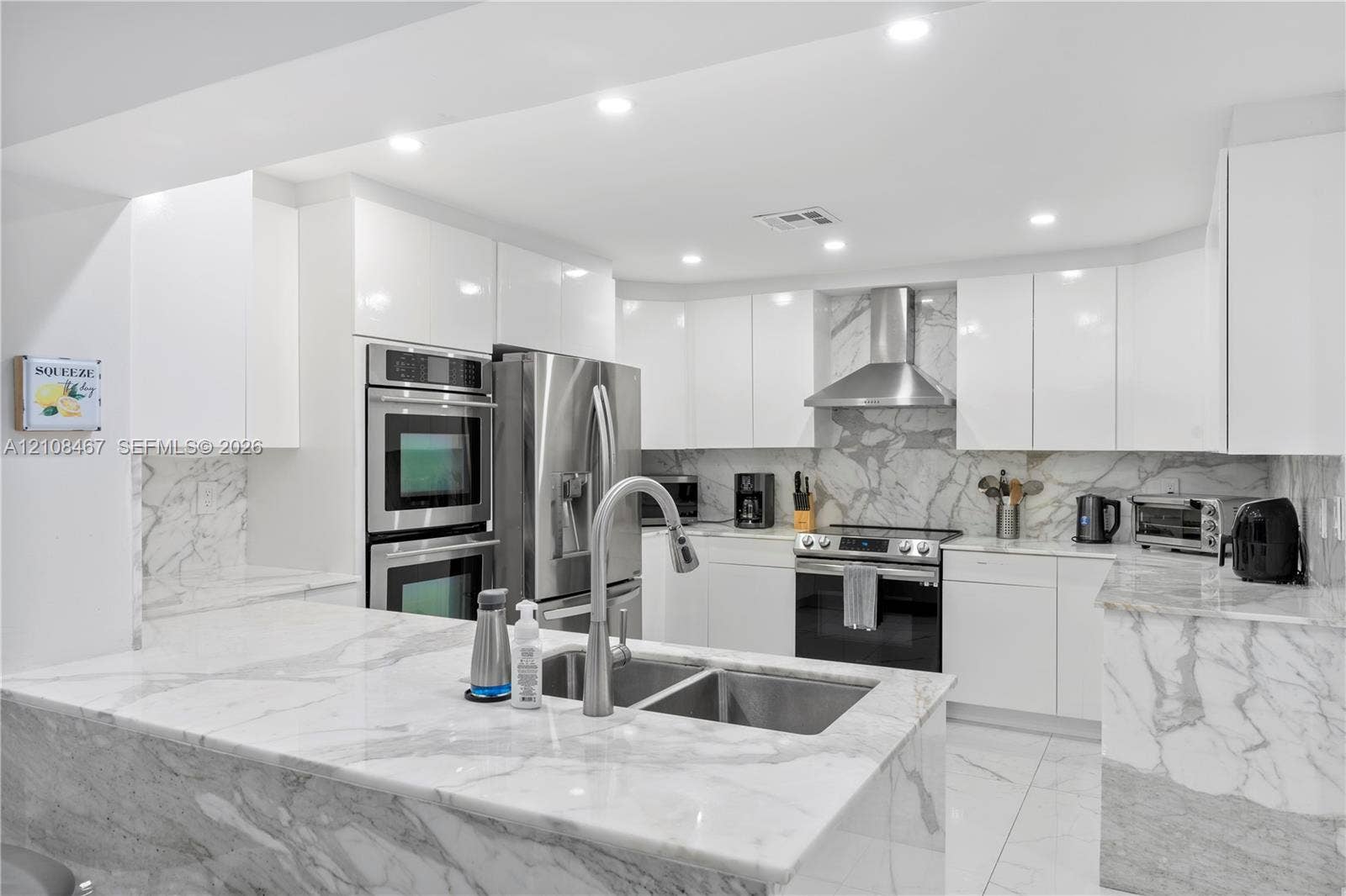 Modern kitchen featuring high-gloss white cabinetry, stone countertops and backsplash, stainless steel built-in double ovens, stainless steel range hood, and recessed lighting