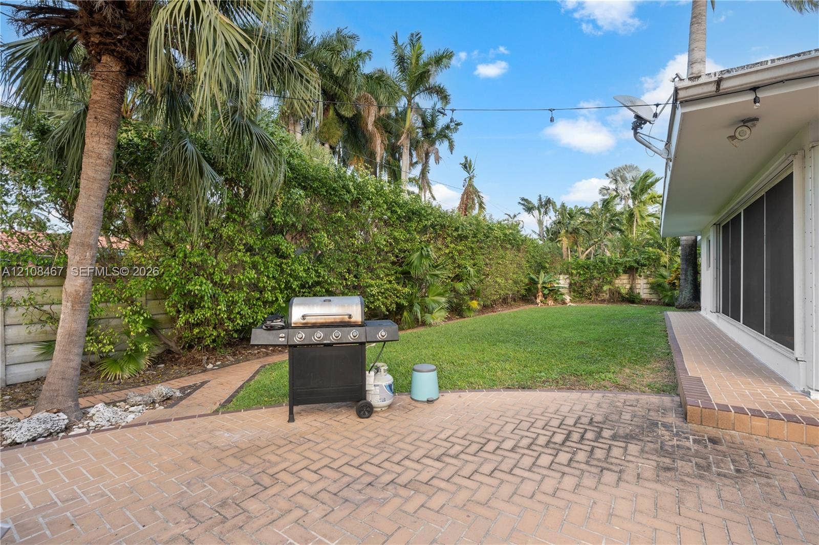 Backyard featuring a herringbone-patterned brick paver patio, established landscaping with mature palm trees, and a green lawn