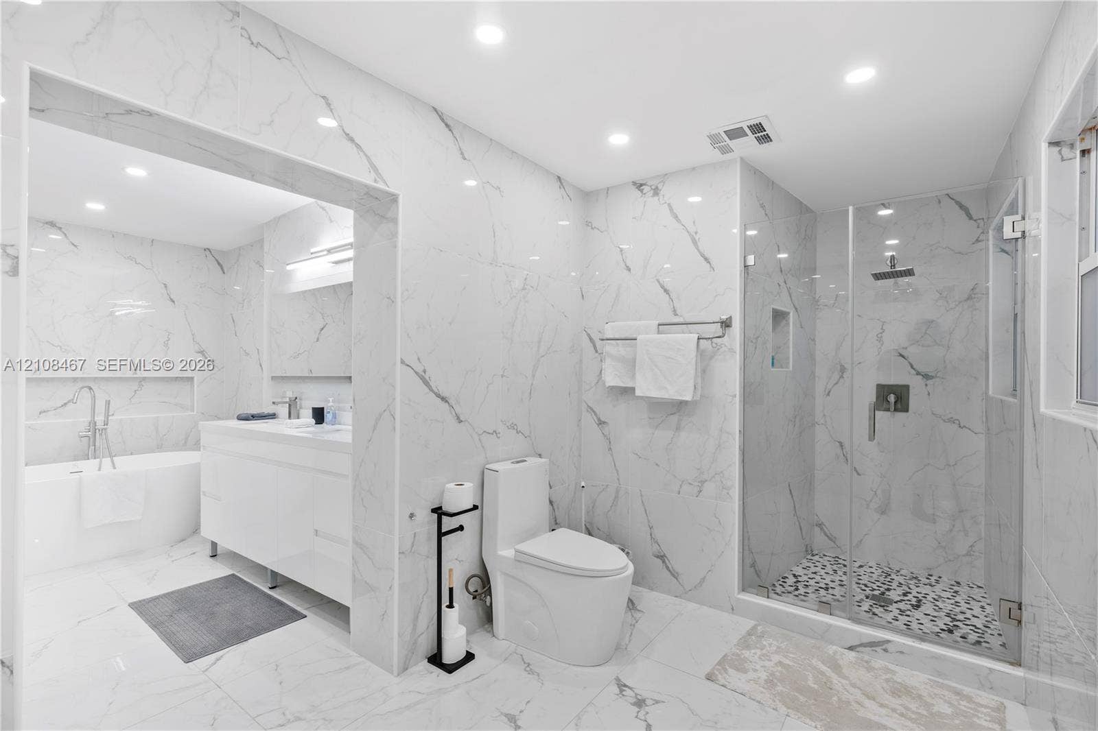 Full-height wall-to-wall stone-finish tile, glass-enclosed shower with pebble-finish flooring, built-in recessed wall niche, freestanding soaking tub, and double-vanity cabinet