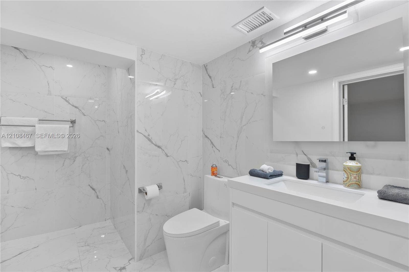 Bathroom featuring floor-to-ceiling large-format tile with marble-pattern finish, vanity with integrated sink and countertop, frameless wall-mounted mirror, overhead LED lighting, and polished chrome fixtures