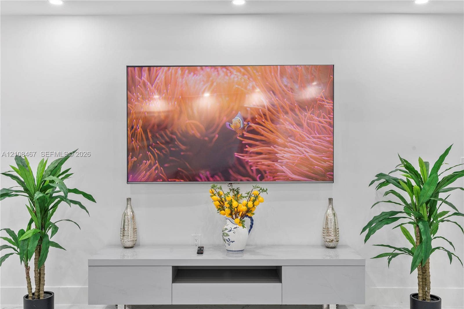 Wall-mounted display screen above a minimalist, light gray media console featuring integrated storage cabinetry and recessed ceiling lighting