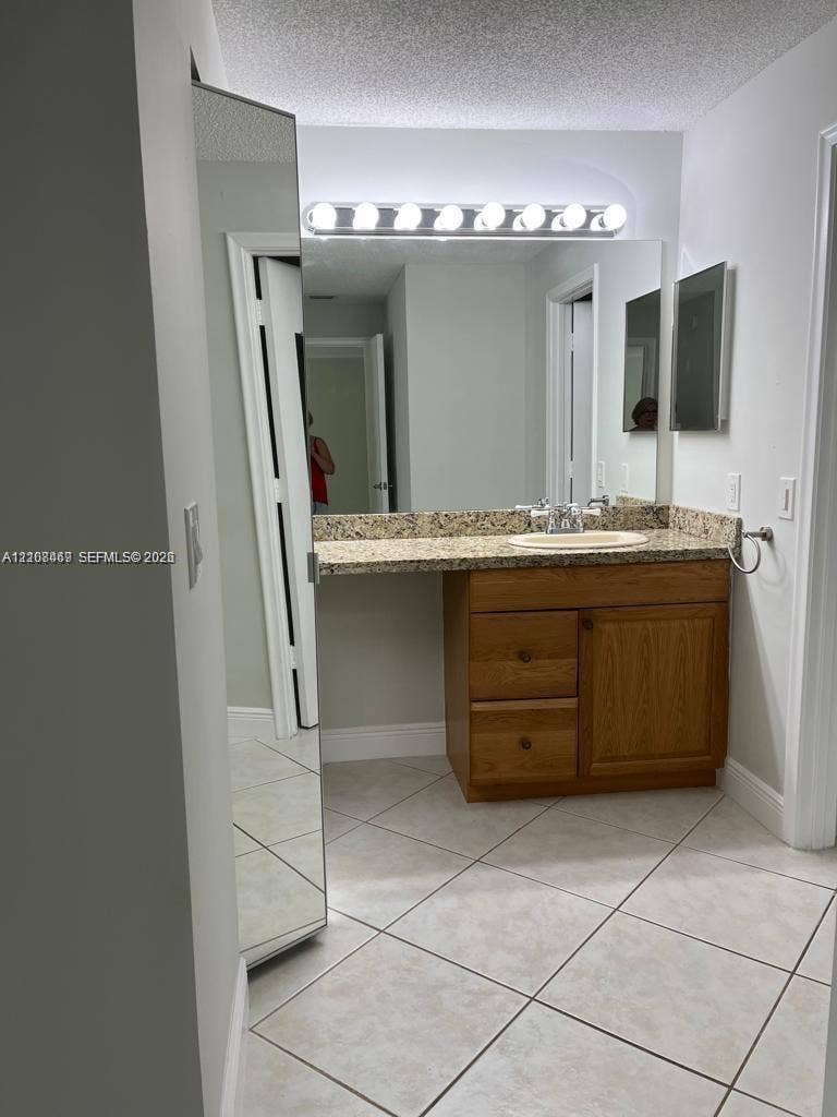 21362 Juego Circle, #33E, Boca Raton, Boca Raton, FL 33433 Vanity featuring wooden cabinetry, stone-finish countertop, oval undermount sink, wall-spanning mirror with integrated light fixture, and diagonal tile flooring.