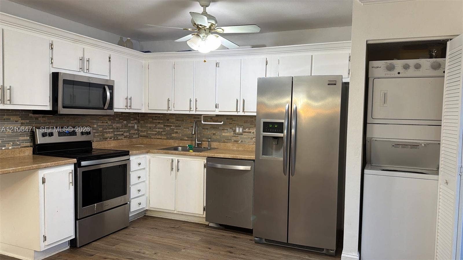 Kitchen featuring white cabinetry, stone-finish countertops, multi-tone tile backsplash, stainless steel appliances, and wood-finish flooring with a stacked laundry unit