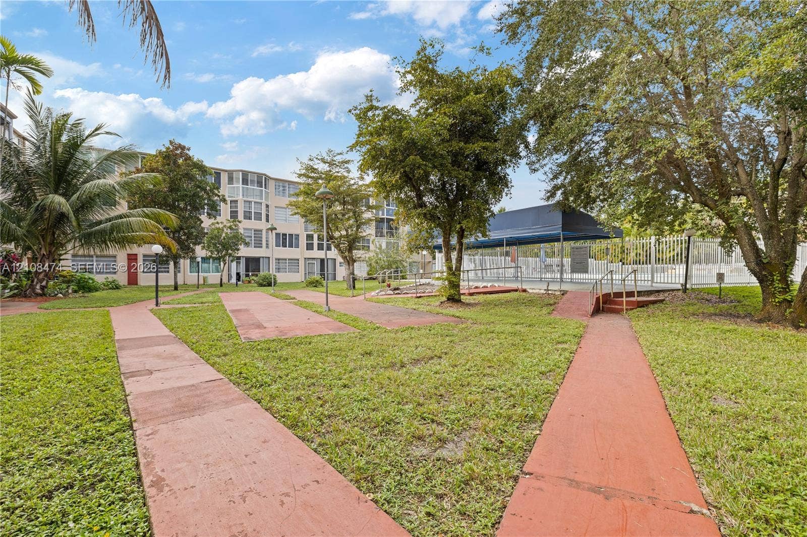 1501 NE 191st Street, #C216, Miami, FL 33179 Common area grounds featuring a landscaped lawn, paved walkways, mature trees, and a shaded pool enclosure