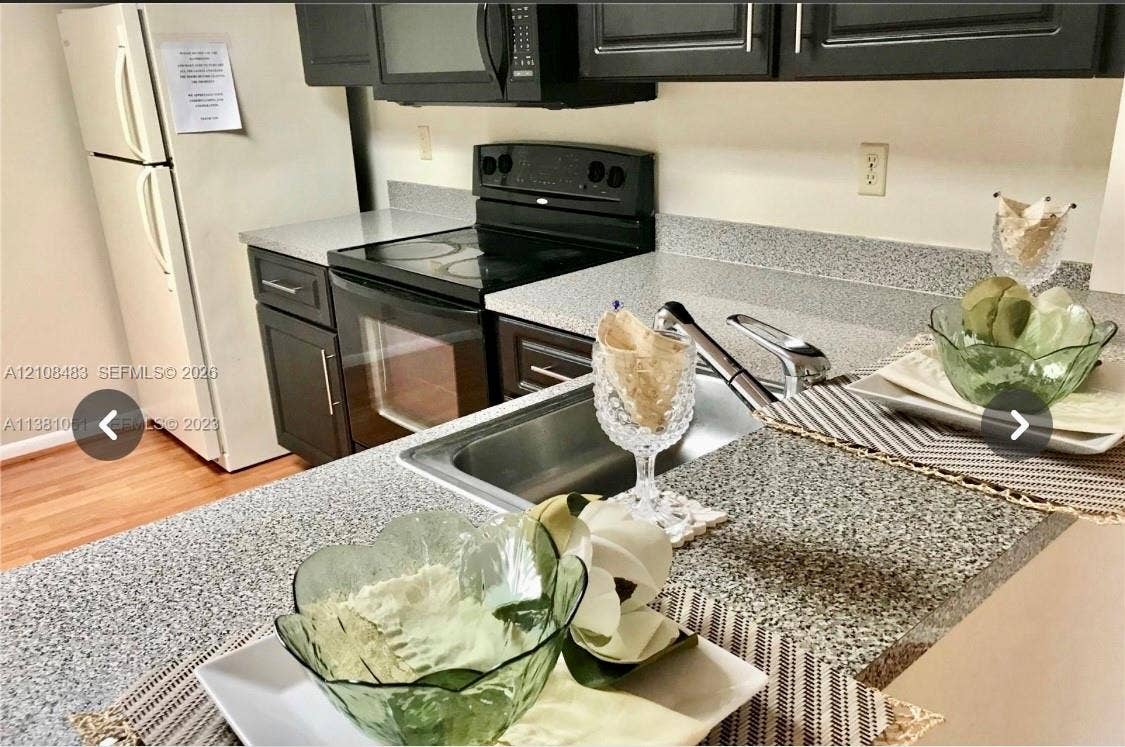 10121 W Sunrise Boulevard, #204, Plantation, Plantation, FL 33322 Kitchen featuring dark cabinetry, speckled stone-finish countertops, stainless steel sink with a high-arch faucet, and black appliances including an electric range and microwave