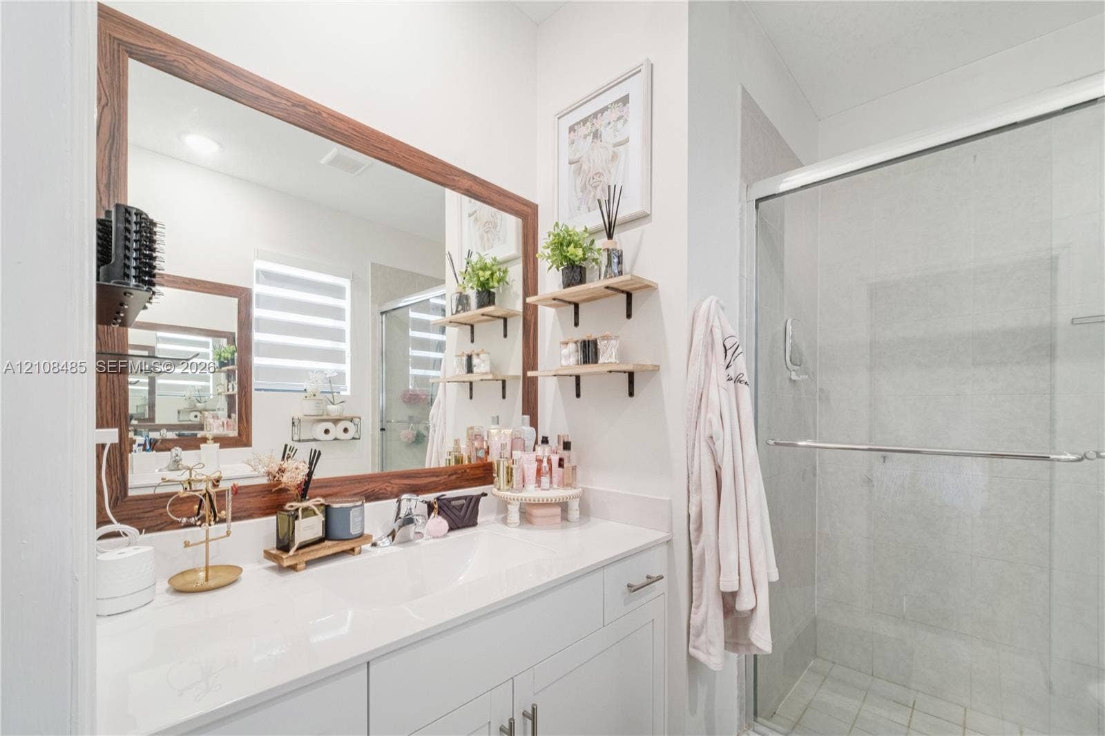 10217 SW 231st Lane, Miami, FL 33190 Vanity featuring a white countertop with an integrated basin, wood-frame mirror, glass-enclosed shower, and wall-mounted shelving