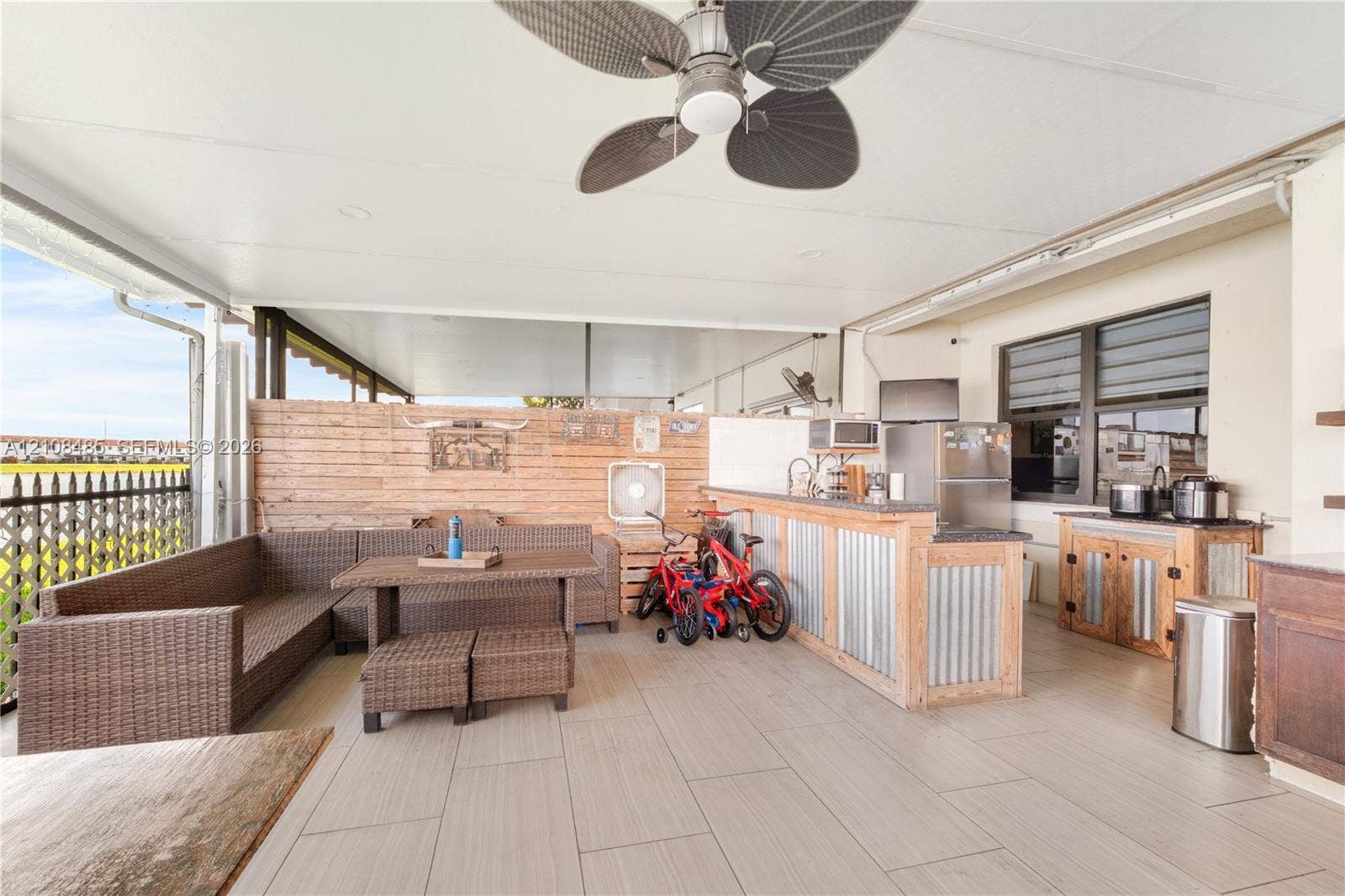 10217 SW 231st Lane, Miami, FL 33190 Covered outdoor patio featuring light-toned tile flooring, built-in bar with metal siding, horizontal wood-slat privacy wall, ceiling fan, and louvered windows