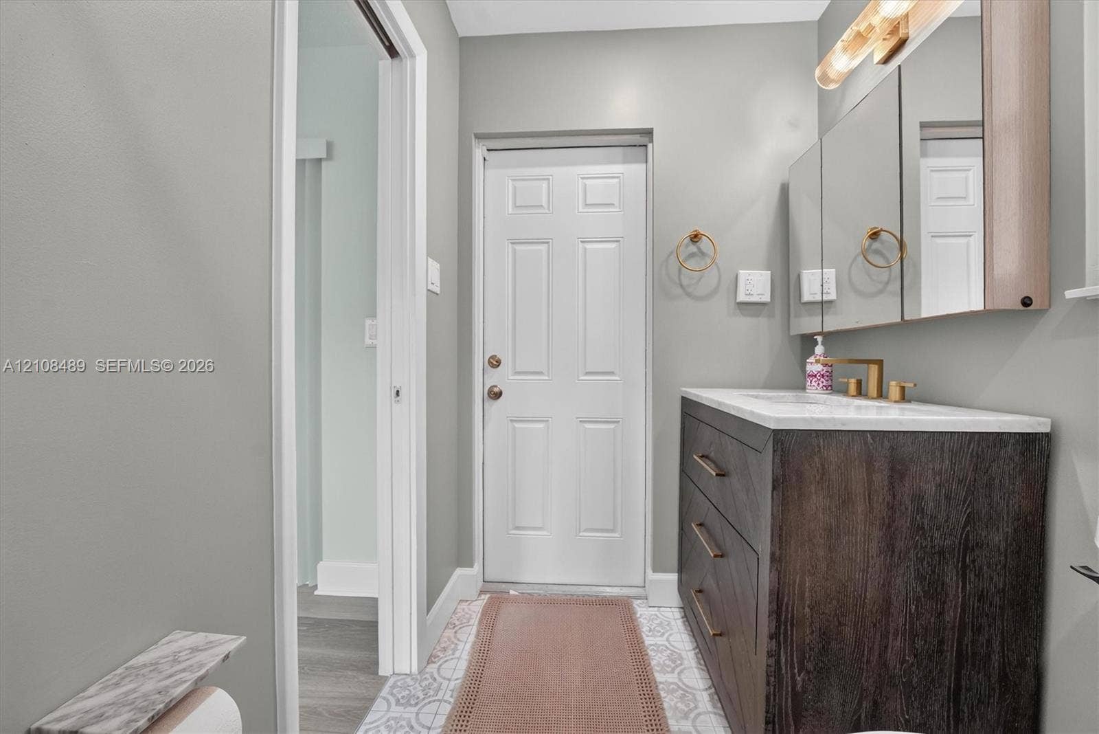Bathroom vanity with dark wood-finish cabinetry, a stone-surface countertop, and gold-tone fixtures, complemented by decorative patterned floor tile and a recessed medicine cabinet