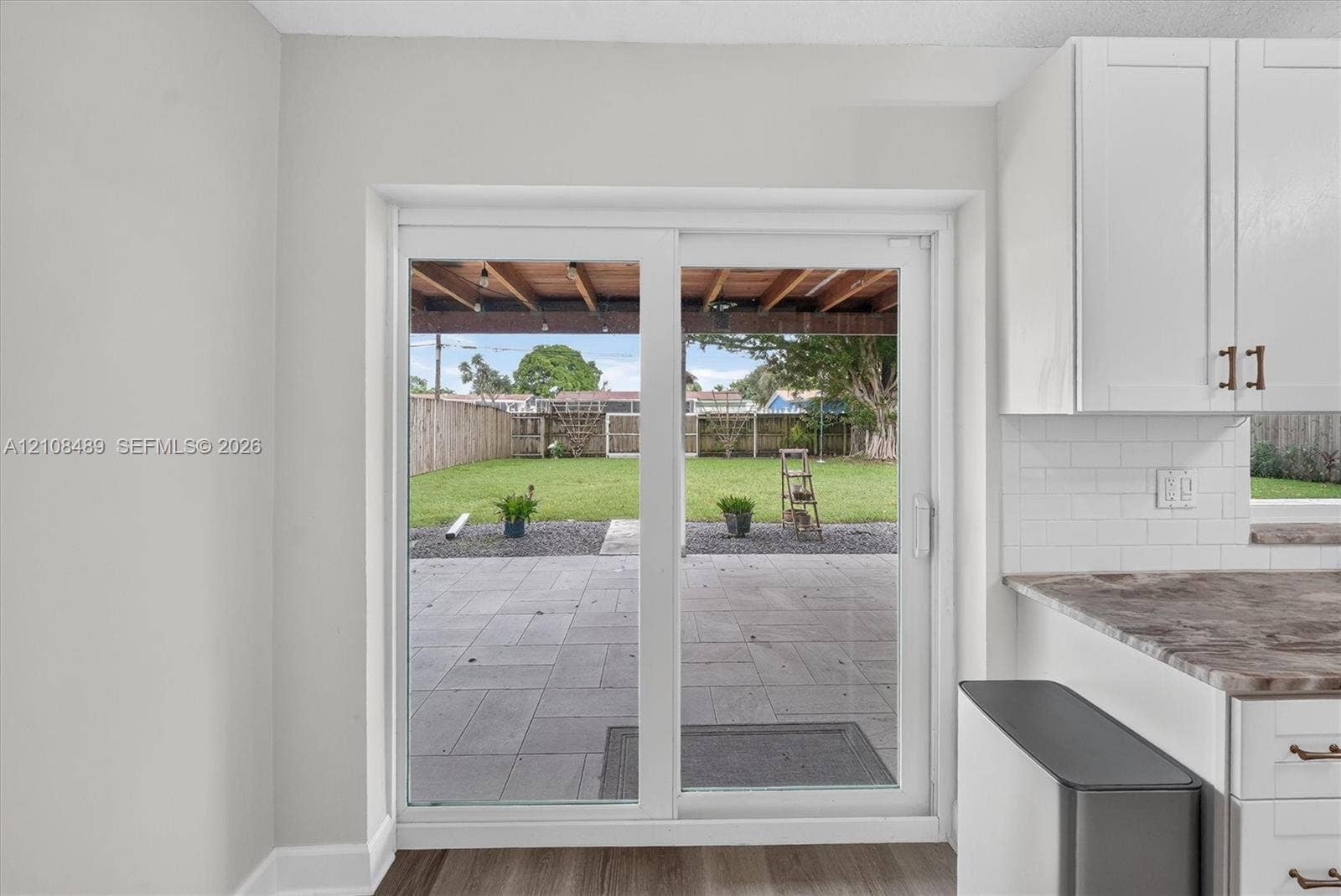 Sliding glass door opening to a concrete patio, covered pergola, fenced backyard with turf, and white shaker-style cabinetry with subway tile backsplash