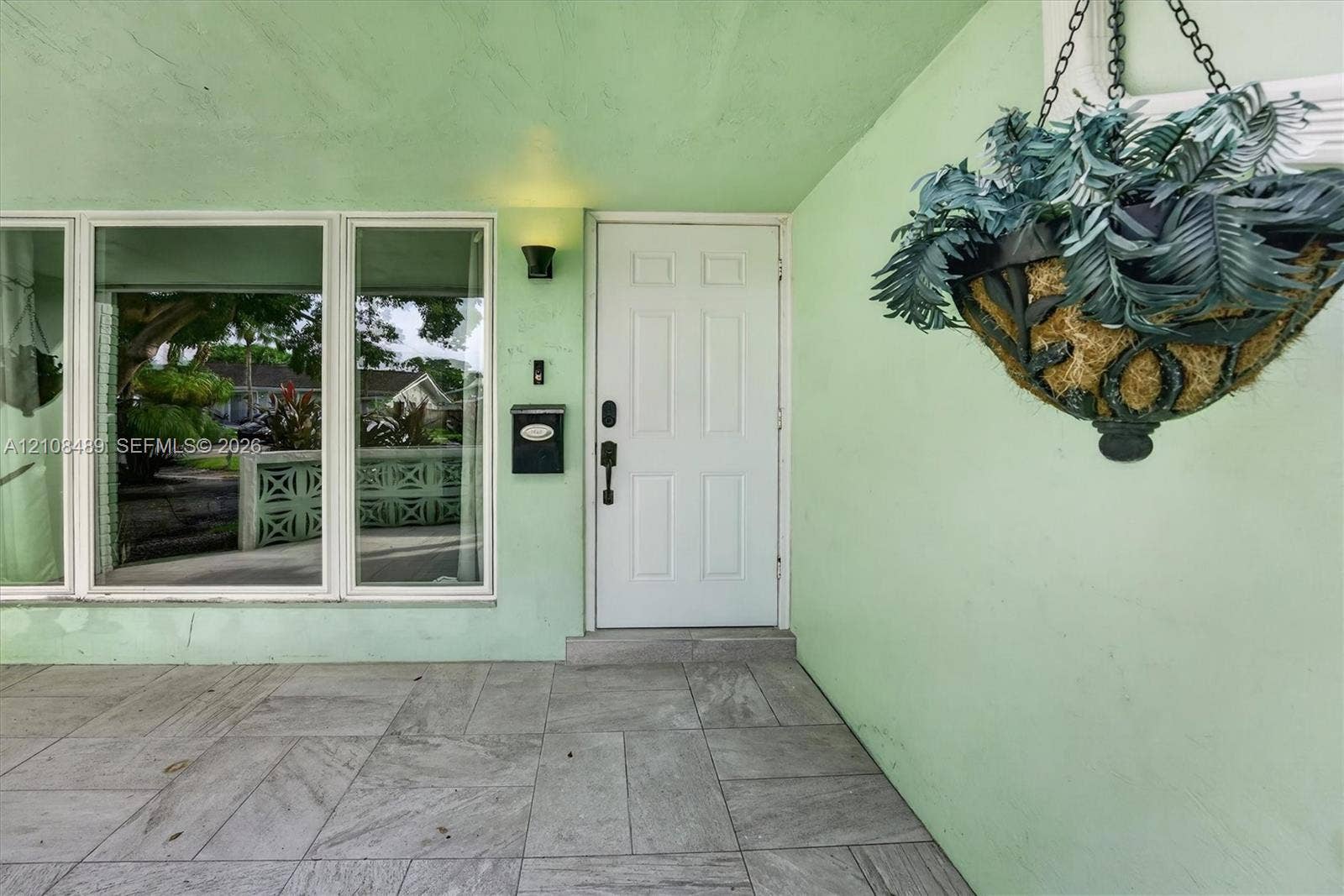 Covered entryway featuring large-format tile flooring, a six-panel exterior door, built-in wall-mounted mailbox, and oversized picture windows