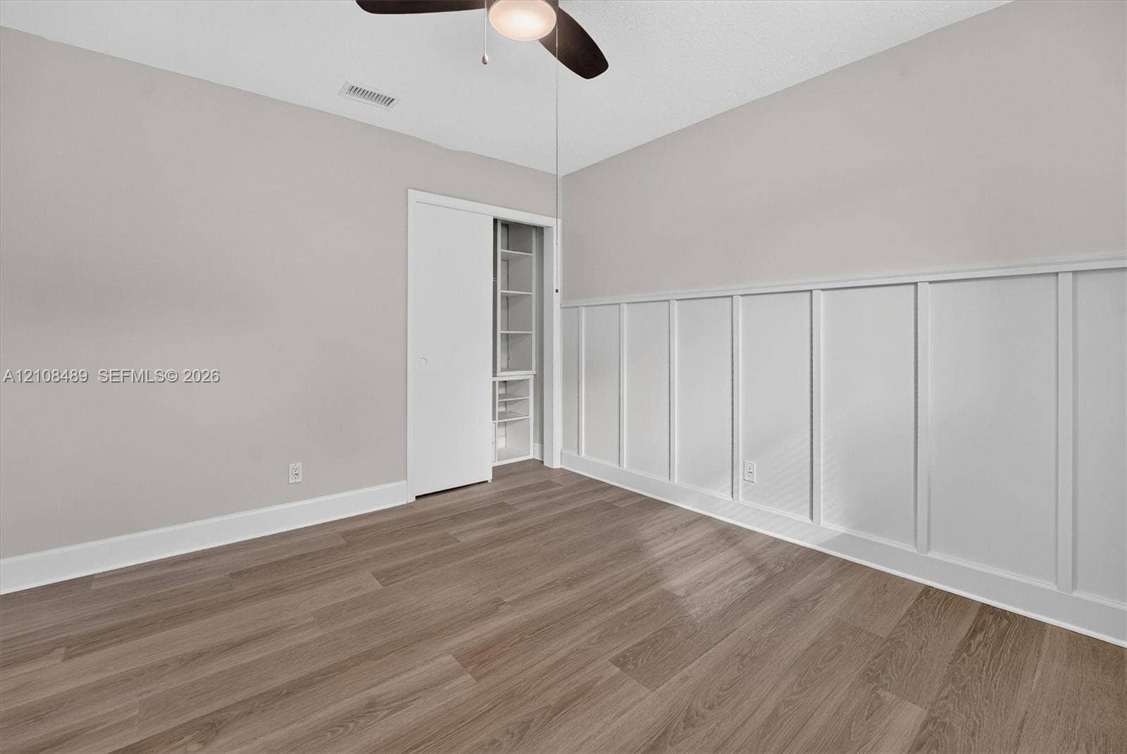 Room featuring white wainscoting, wood-finish flooring, a ceiling-mounted light fixture with a fan, and a closet with built-in shelving