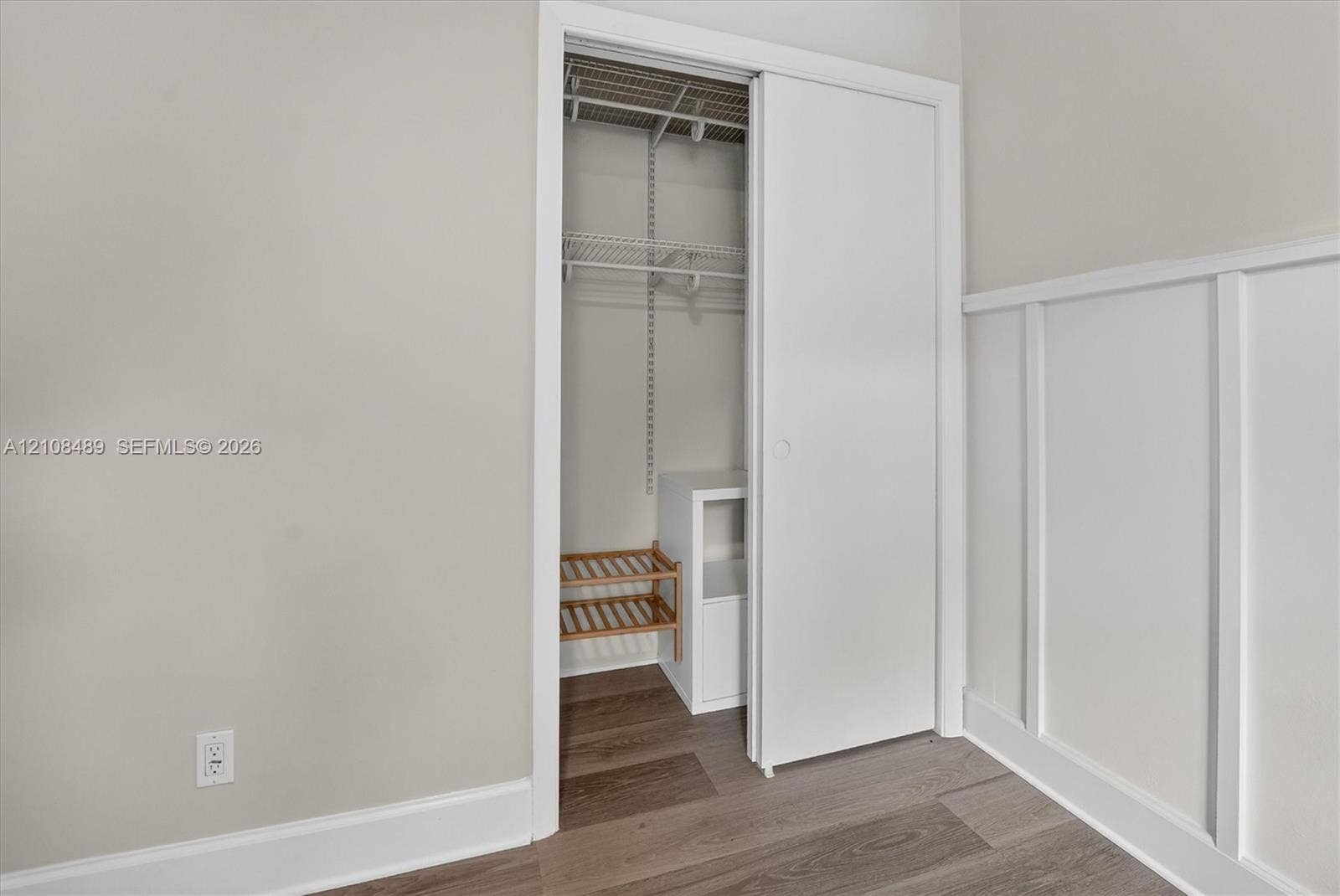 Closet with white sliding door, adjustable wire shelving, and wood-finish flooring, complemented by wall-mounted wainscoting
