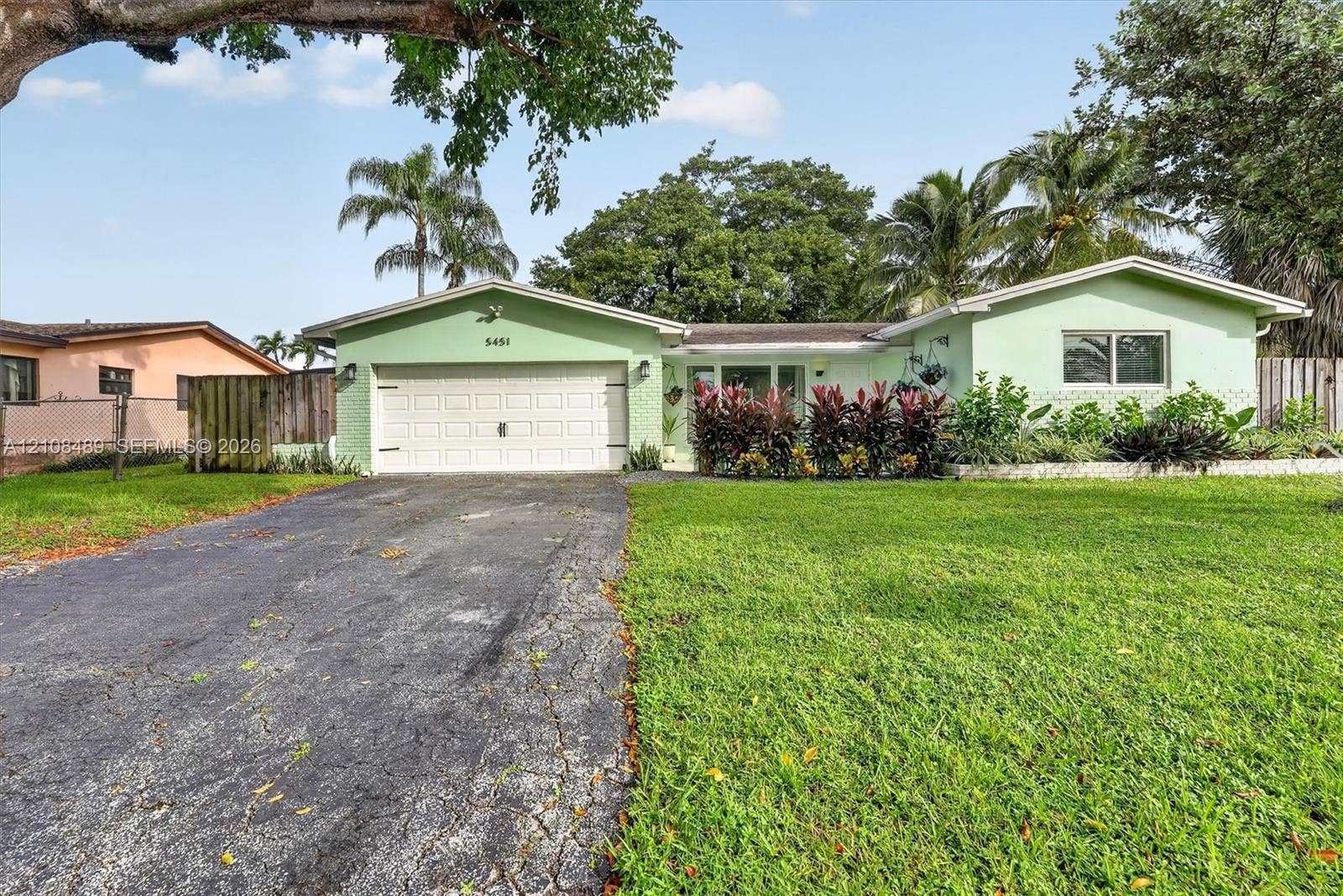 Single-story residence featuring a painted masonry exterior, a wide asphalt driveway, an attached two-car garage, and established landscaping with mature palms