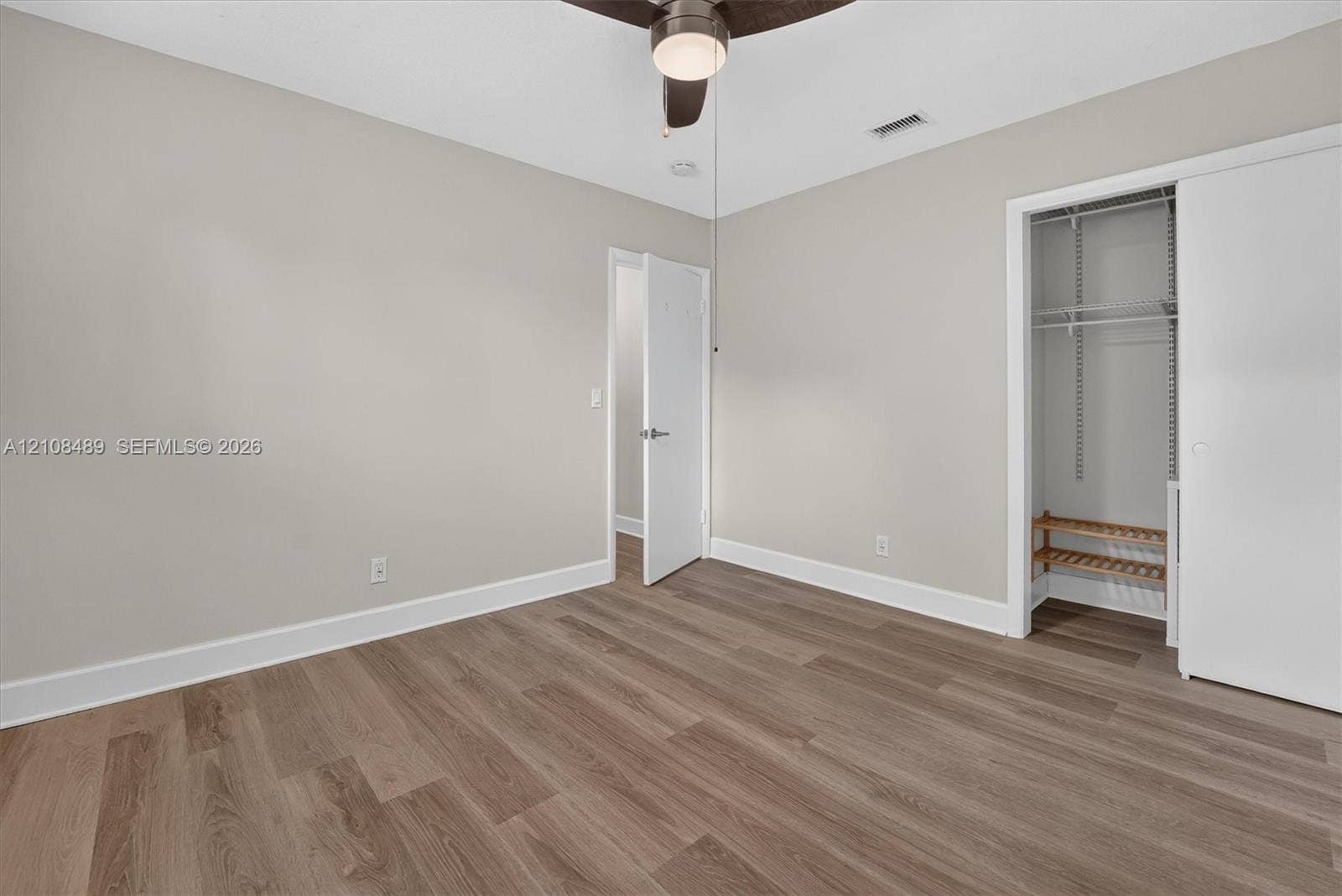 Neutral-toned interior space featuring wood-finish flooring, a ceiling fan with integrated lighting, and a closet with built-in wire shelving