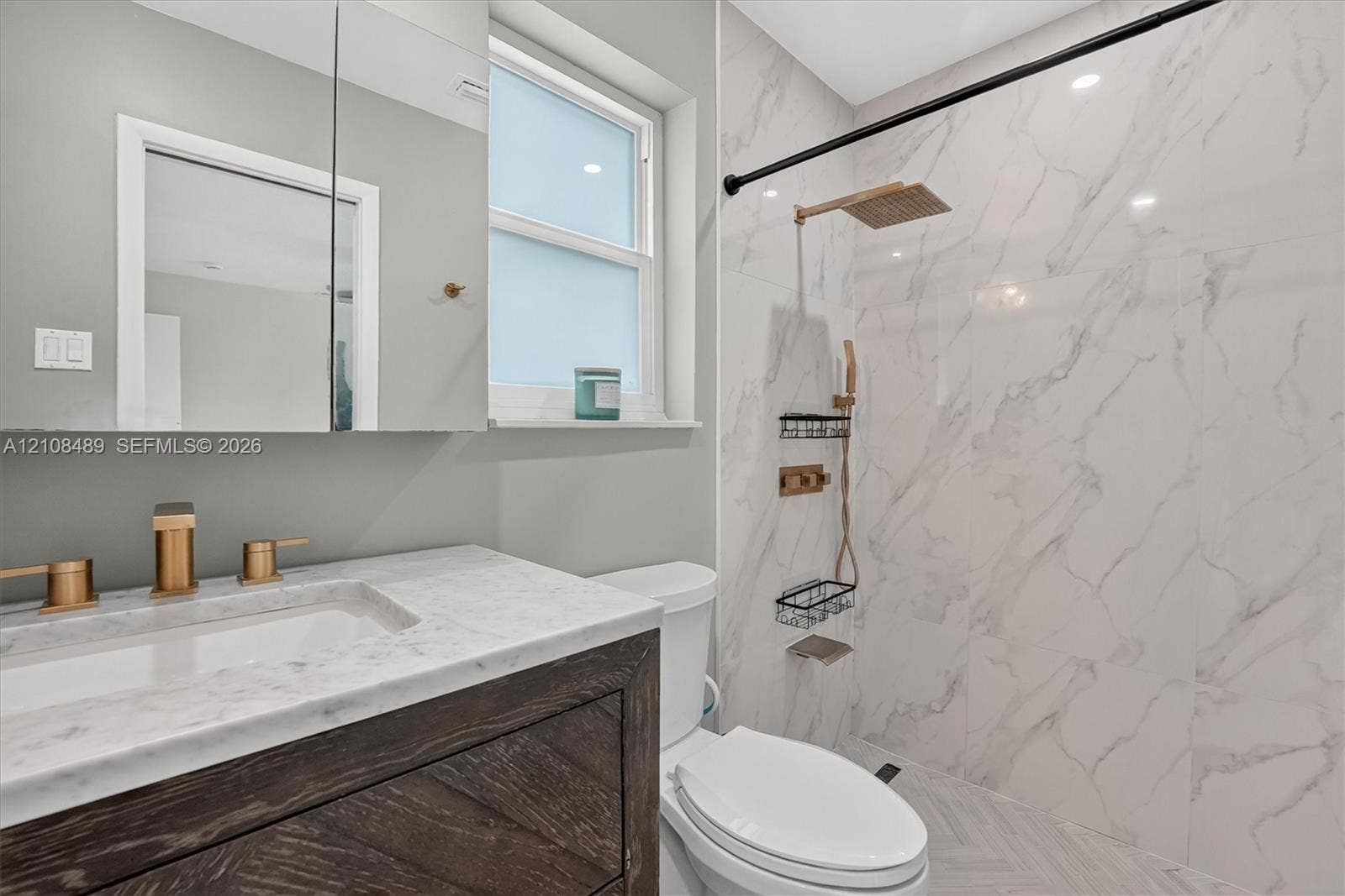 Bathroom featuring a vanity with wood-finish cabinetry, a light-toned stone countertop, brass-finish fixtures, a shower enclosure with large-format stone-patterned wall tile, and herringbone-patterned floor tile