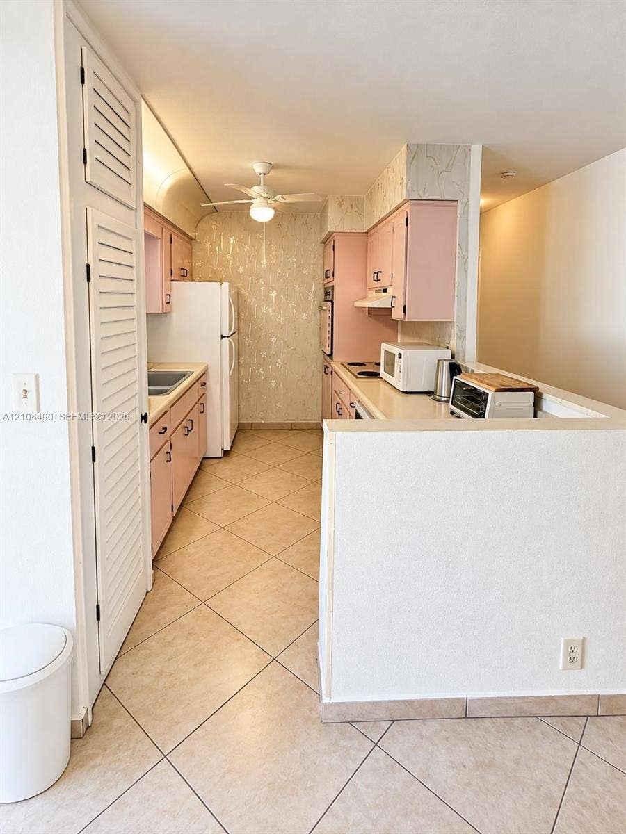 Kitchen featuring cabinetry with integrated hardware, neutral tile flooring, built-in pantry with louvered doors, and a ceiling-mounted light fixture with fan