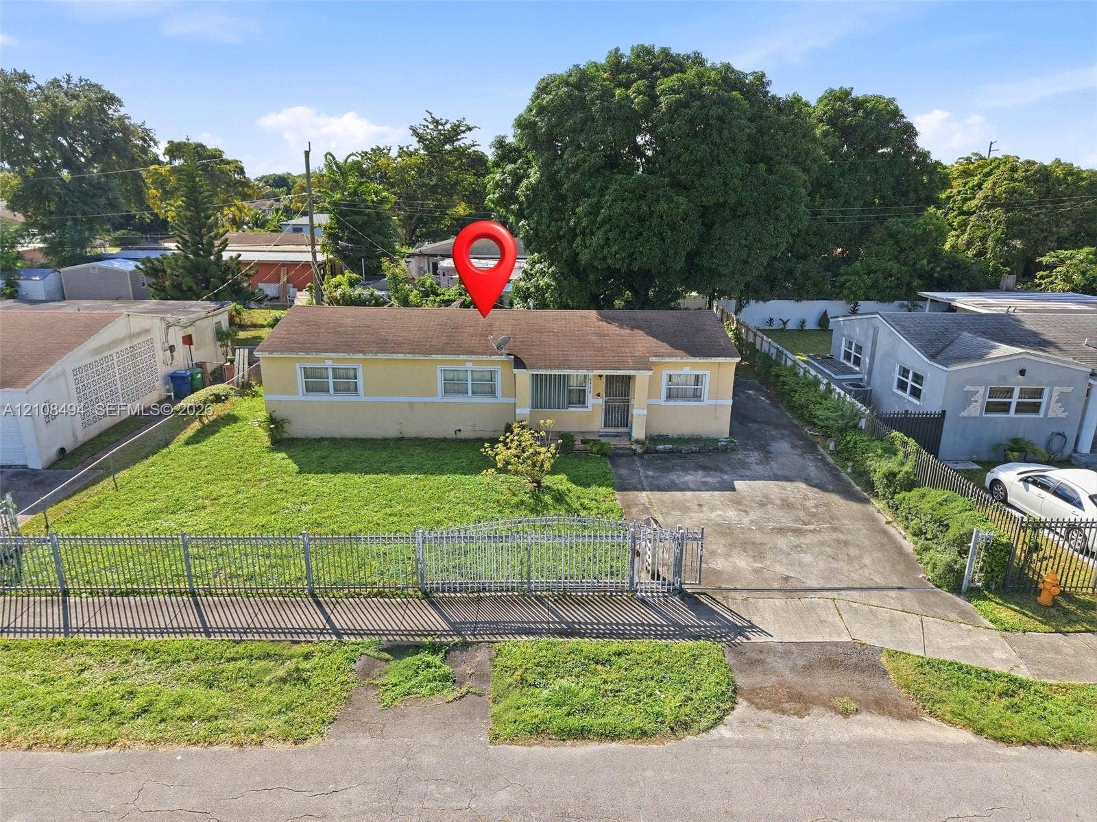 6940 NW 28th Avenue, Miami, FL 33147 Single-story residence featuring a low-slope roof, stucco exterior, paved driveway, metal perimeter fencing, and mature trees