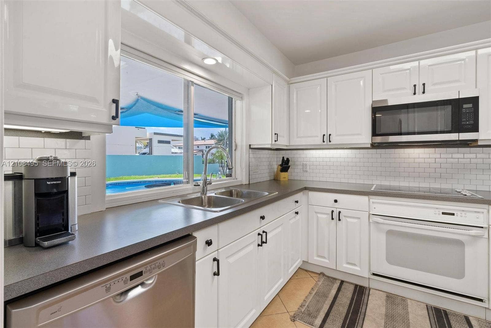 3080 NE 43rd Street, Fort Lauderdale, FL 33308 Kitchen featuring white shaker-style cabinetry, solid-surface countertops, a subway tile backsplash, and a stainless steel dishwasher and microwave