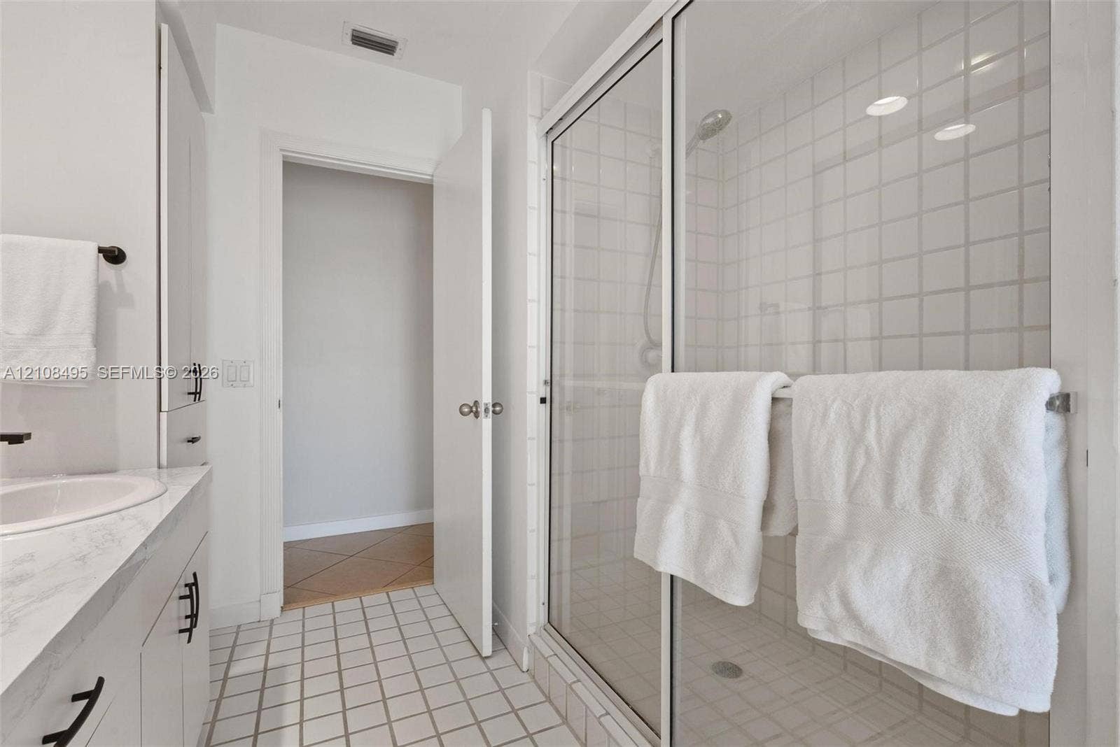 3080 NE 43rd Street, Fort Lauderdale, FL 33308 Bathroom featuring a vanity with a white countertop and integrated sink, a glass-enclosed shower with tiled surround, and a tall built-in storage cabinet with matte black hardware