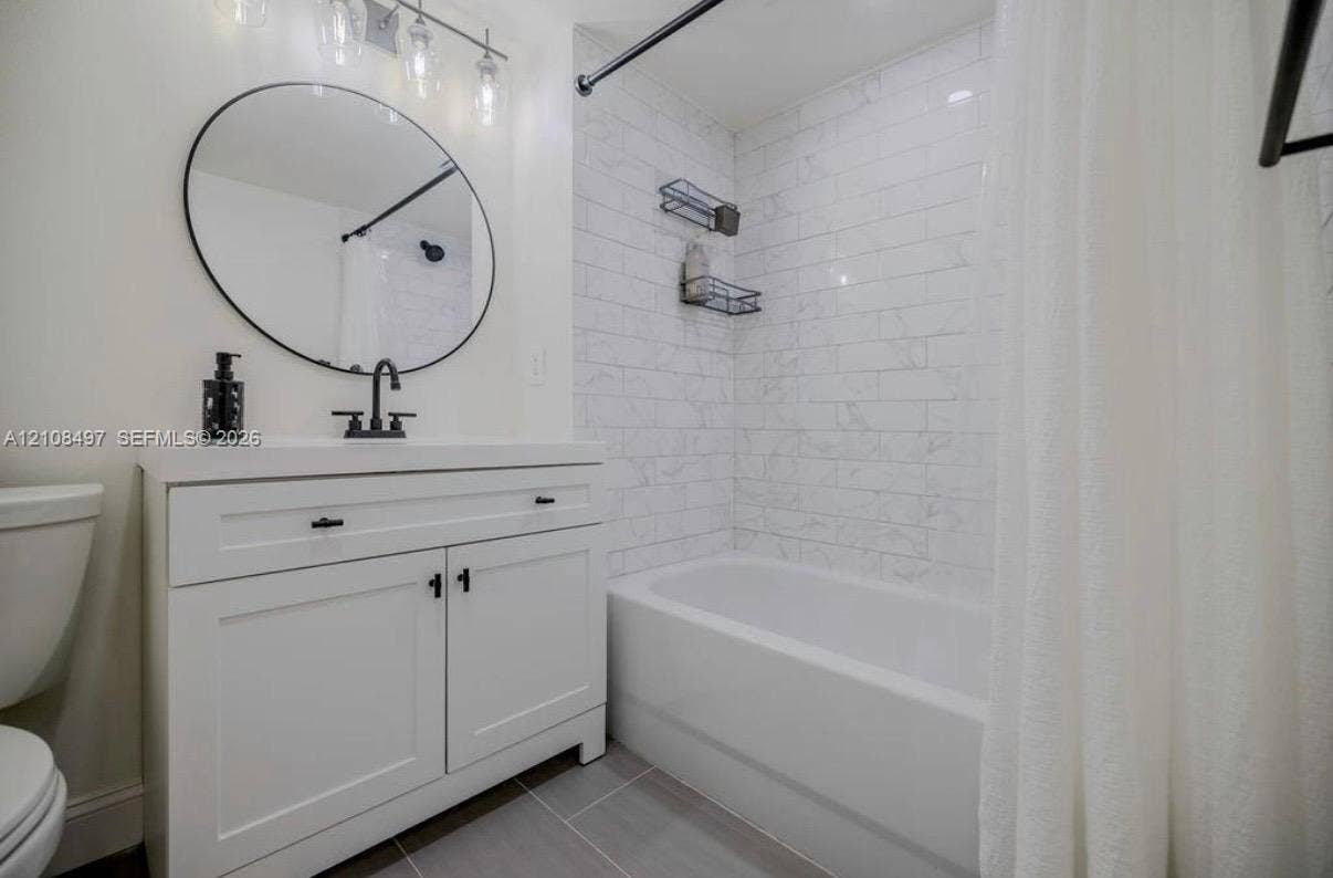 6245 SW 47th Manor, #101, Davie, FL 33314 White vanity with shaker-style cabinetry, matte black faucet, round wall-mounted mirror, light-toned subway tile shower surround, and grey tile flooring